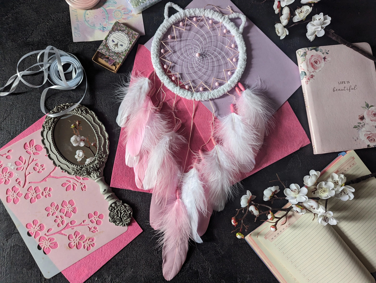 Handcrafted pink and white dream catcher with a cat ear design, featuring a pink beaded web and delicate white and pink feathers. This bohemian decor piece is surrounded by crafting materials and floral elements, making it an ideal gift for a nursery or girl's room. The soft pastel colors and intricate detailing add a touch of whimsy and charm, perfect for hanging in a bedroom or as a unique decorative accent in any space. Enhance your home decor with this adorable, handmade dream catcher.