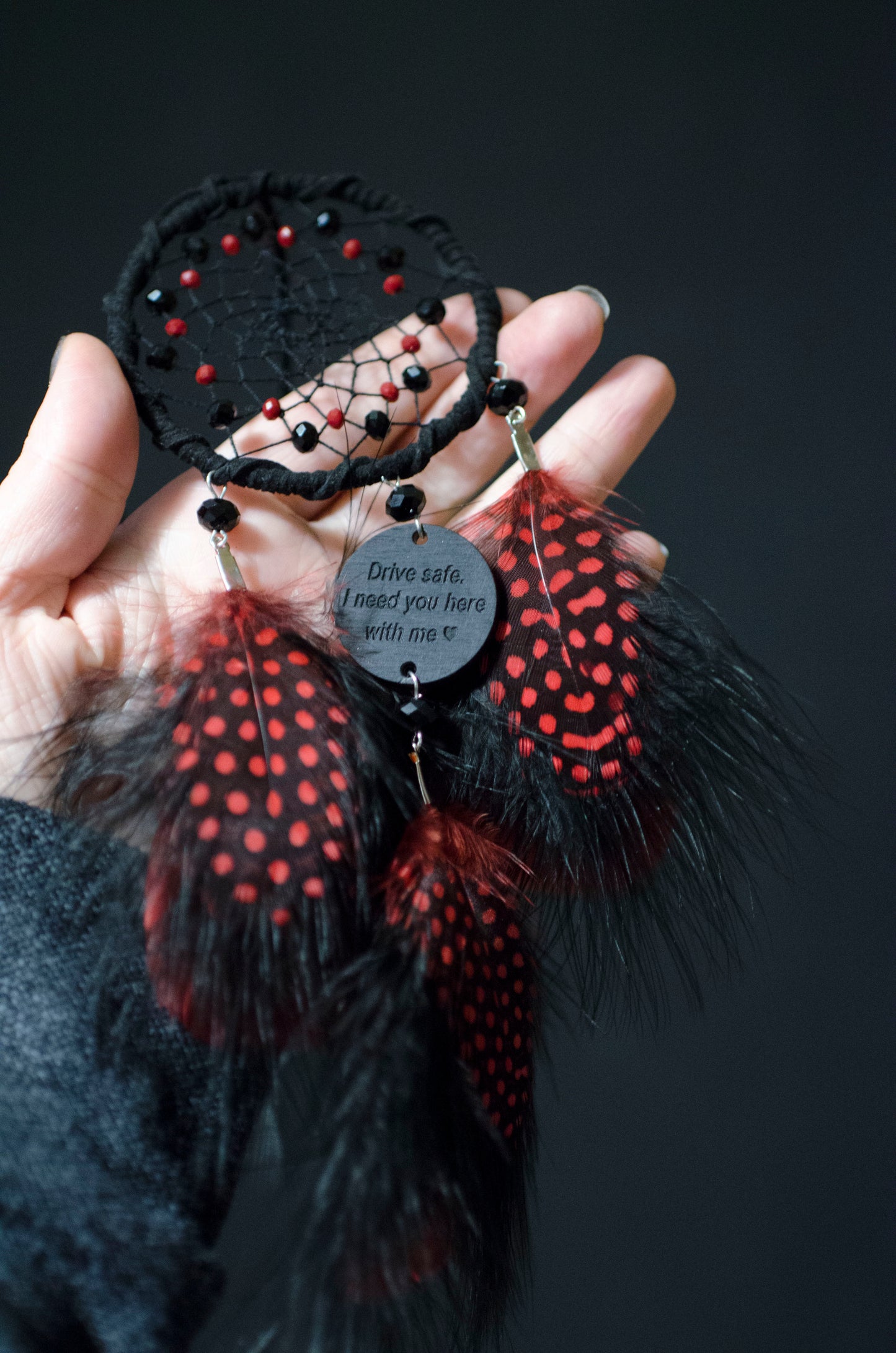 Handcrafted black and red dream catcher with vibrant feathers, designed for car rearview mirrors. Features an inspirational message charm, "Drive safe. I need you here with me." Perfect for adding a touch of protection and style to your car interior.