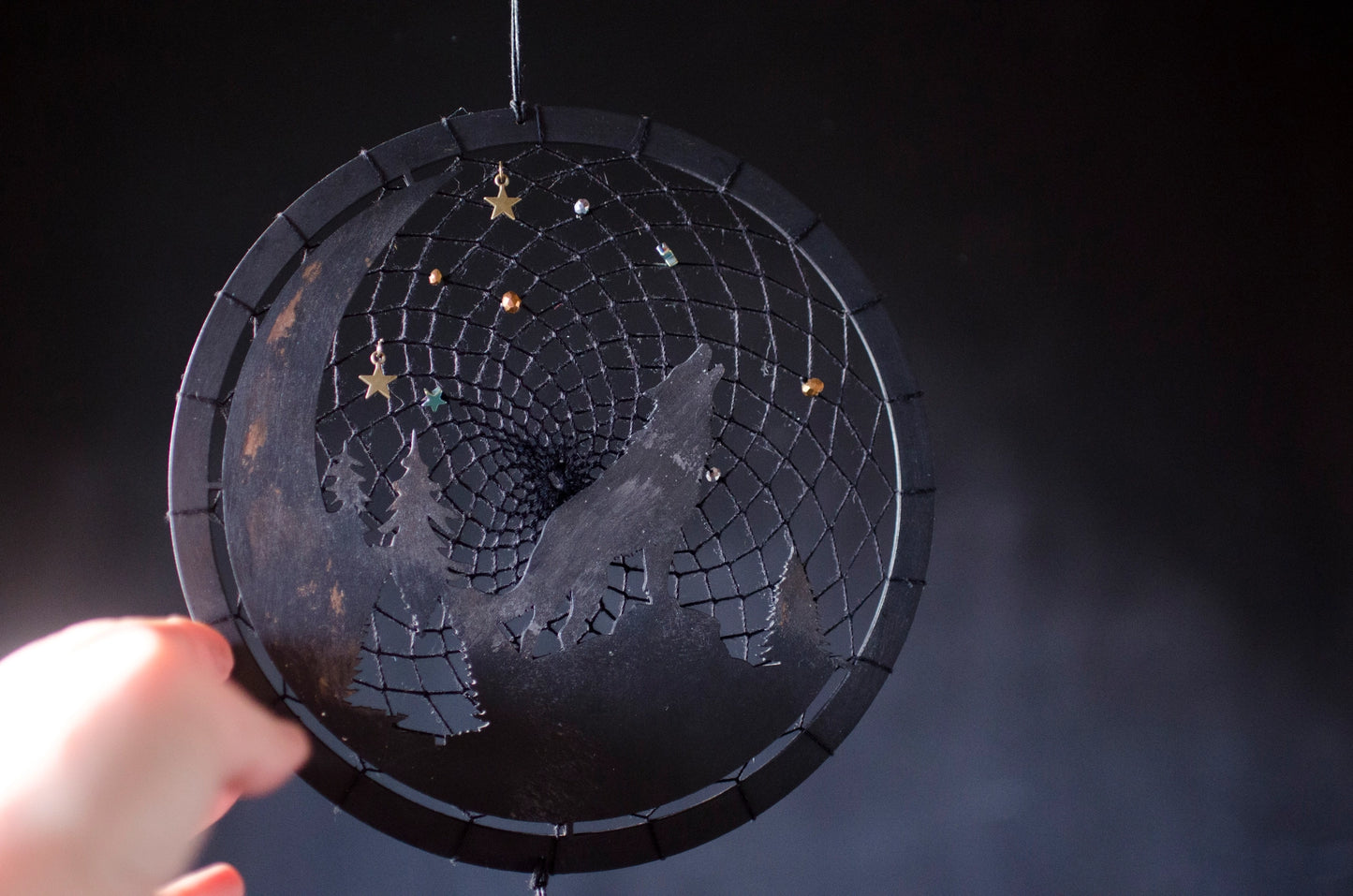 A handcrafted dream catcher with a howling wolf silhouette, moon, and forest design. Decorated with star-shaped beads, this unique piece captures the essence of the night. The hoop measures approximately 8 inches in diameter, featuring black and natural shades.