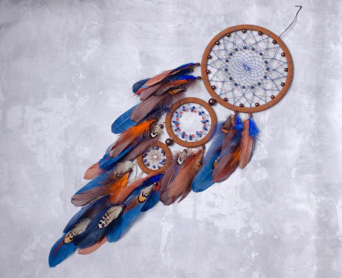 Handcrafted triple hoop dreamcatcher with brown wooden rings, blue and brown feathers, and beaded web, hanging on a wall surrounded by indoor plants in natural sunlight.