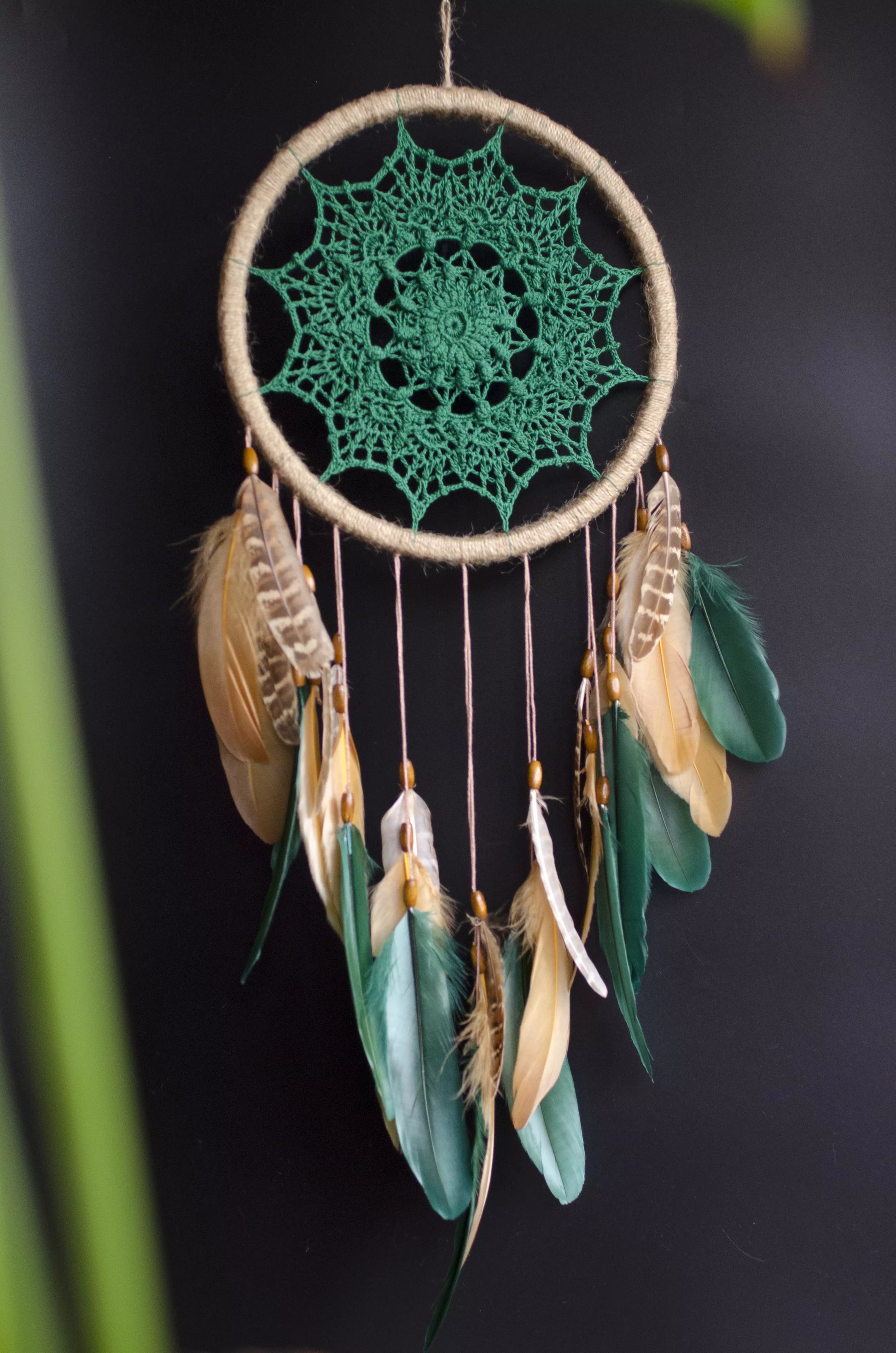 A handcrafted dreamcatcher featuring a green crochet mandala centerpiece, wrapped in a jute hoop with natural brown and green feathers hanging below, styled against dark and light backgrounds for a modern, bohemian look.