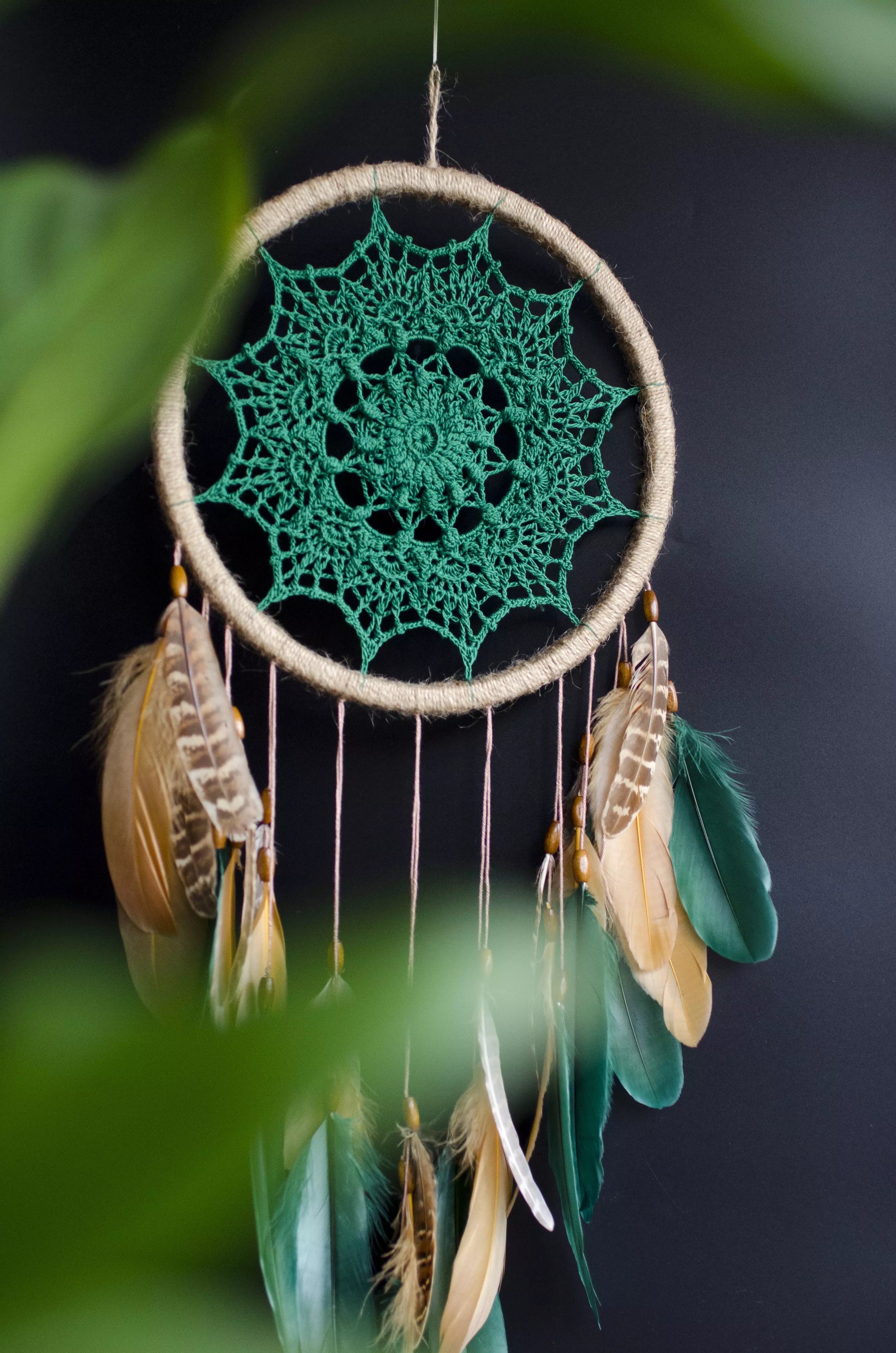 A handcrafted dreamcatcher featuring a green crochet mandala centerpiece, wrapped in a jute hoop with natural brown and green feathers hanging below, styled against dark and light backgrounds for a modern, bohemian look.