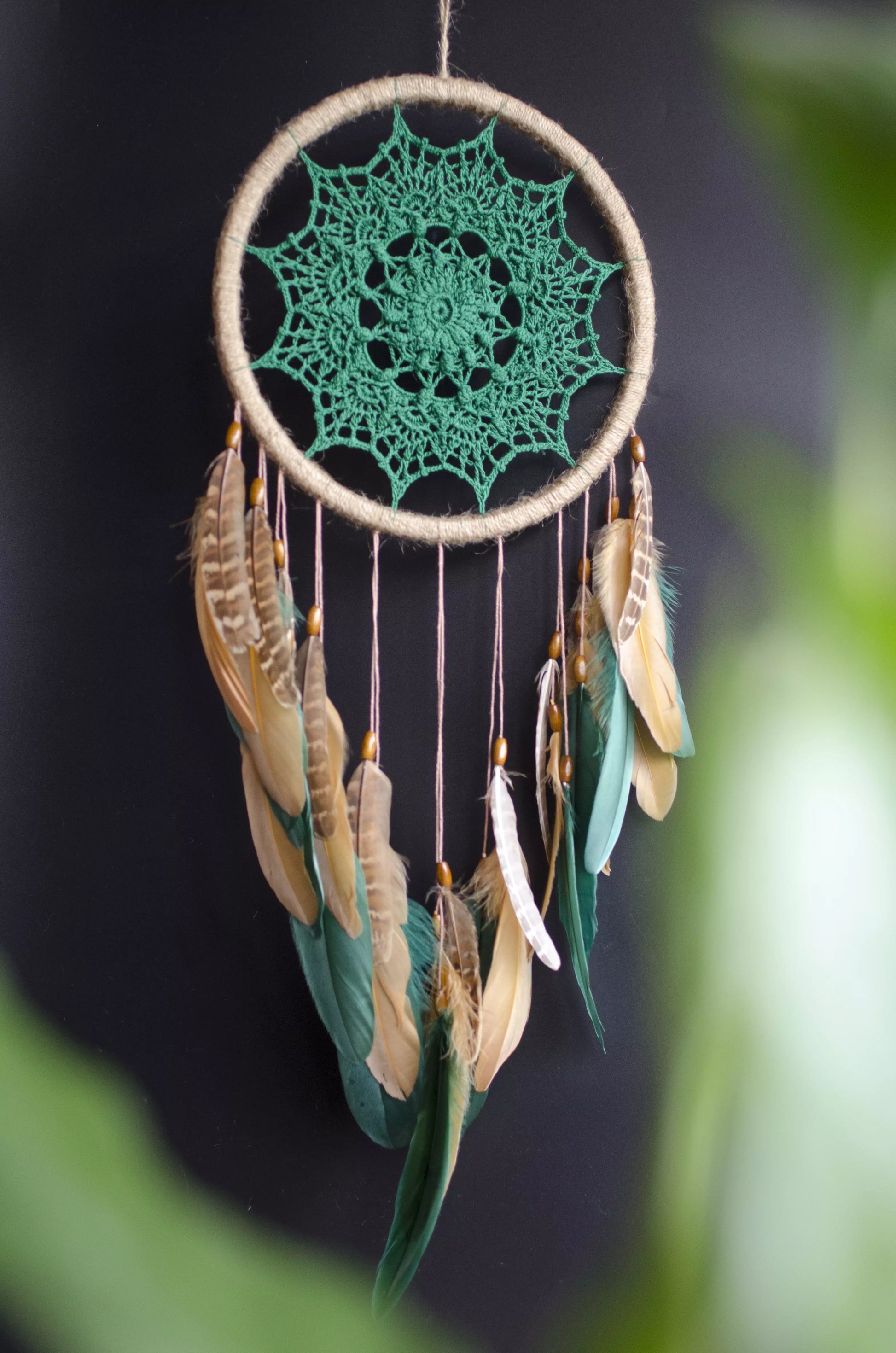 A handcrafted dreamcatcher featuring a green crochet mandala centerpiece, wrapped in a jute hoop with natural brown and green feathers hanging below, styled against dark and light backgrounds for a modern, bohemian look.