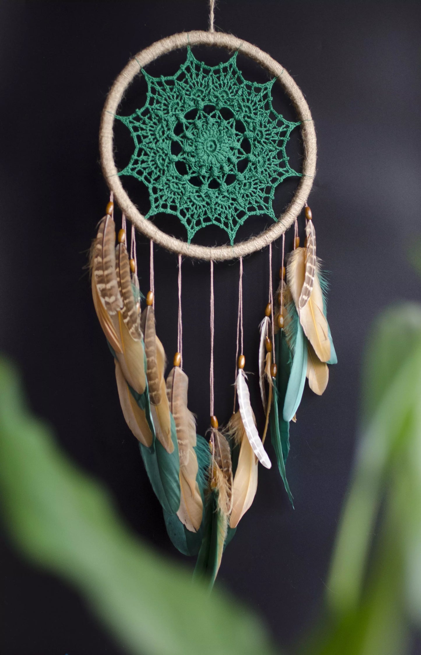 A handcrafted dreamcatcher featuring a green crochet mandala centerpiece, wrapped in a jute hoop with natural brown and green feathers hanging below, styled against dark and light backgrounds for a modern, bohemian look.