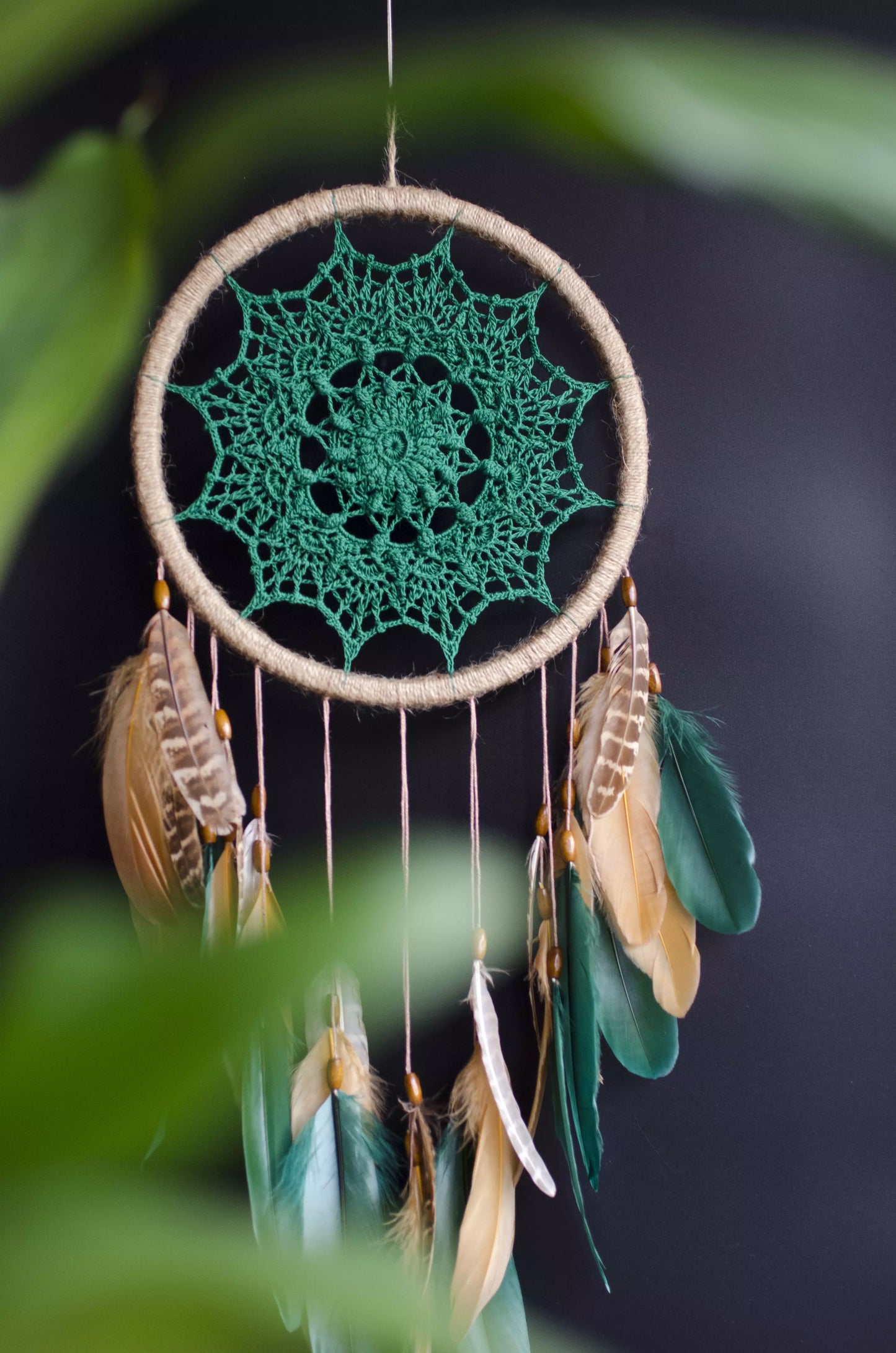 A handcrafted dreamcatcher featuring a green crochet mandala centerpiece, wrapped in a jute hoop with natural brown and green feathers hanging below, styled against dark and light backgrounds for a modern, bohemian look.