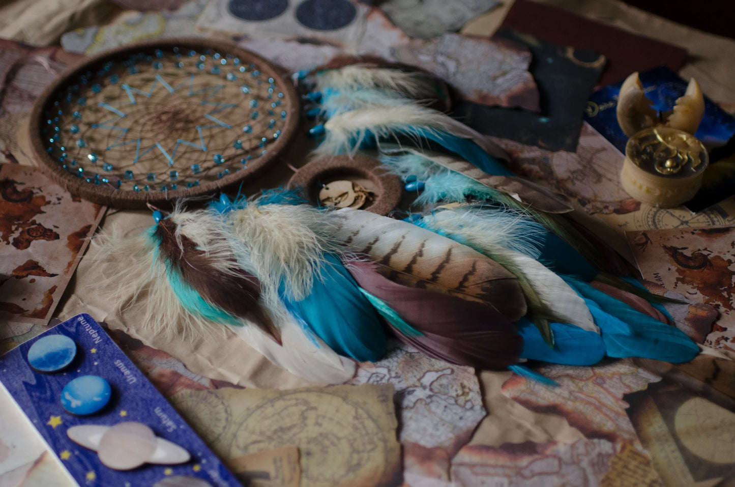 Handcrafted dream catcher with a large brown hoop featuring an intricate web adorned with blue beads. Decorated with natural feathers in shades of blue, brown, and white, it includes a central wooden charm with an engraved design. The dream catcher is set against a backdrop of vintage maps and celestial charts, creating a mystical and adventurous aesthetic. Perfect for adding a bohemian touch to any space, it symbolizes protection and positive energy, making it an ideal gift or home decor piece for dreamers