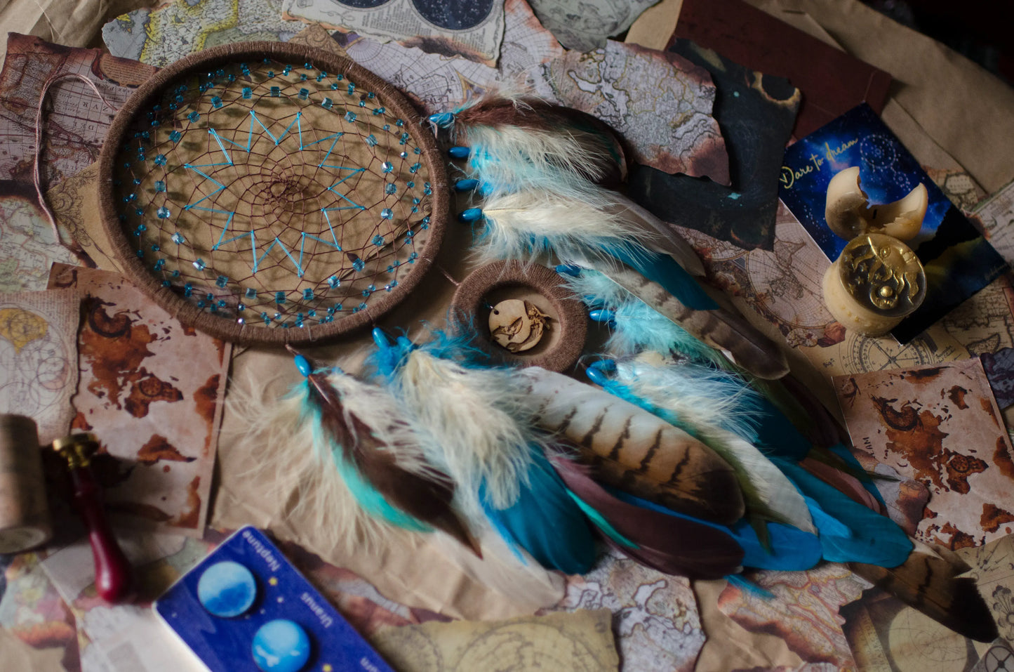 Handcrafted dream catcher with a large brown hoop featuring an intricate web adorned with blue beads. Decorated with natural feathers in shades of blue, brown, and white, it includes a central wooden charm with an engraved design. The dream catcher is set against a backdrop of vintage maps and celestial charts, creating a mystical and adventurous aesthetic. Perfect for adding a bohemian touch to any space, it symbolizes protection and positive energy, making it an ideal gift or home decor piece for dreamers