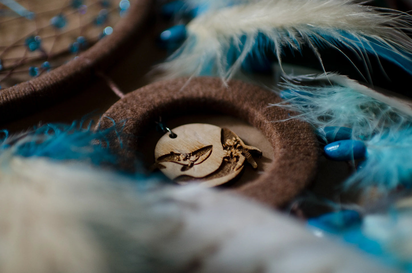 Handcrafted dream catcher with a large brown hoop featuring an intricate web adorned with blue beads. Decorated with natural feathers in shades of blue, brown, and white, it includes a central wooden charm with an engraved design. The dream catcher is set against a backdrop of vintage maps and celestial charts, creating a mystical and adventurous aesthetic. Perfect for adding a bohemian touch to any space, it symbolizes protection and positive energy, making it an ideal gift or home decor piece for dreamers