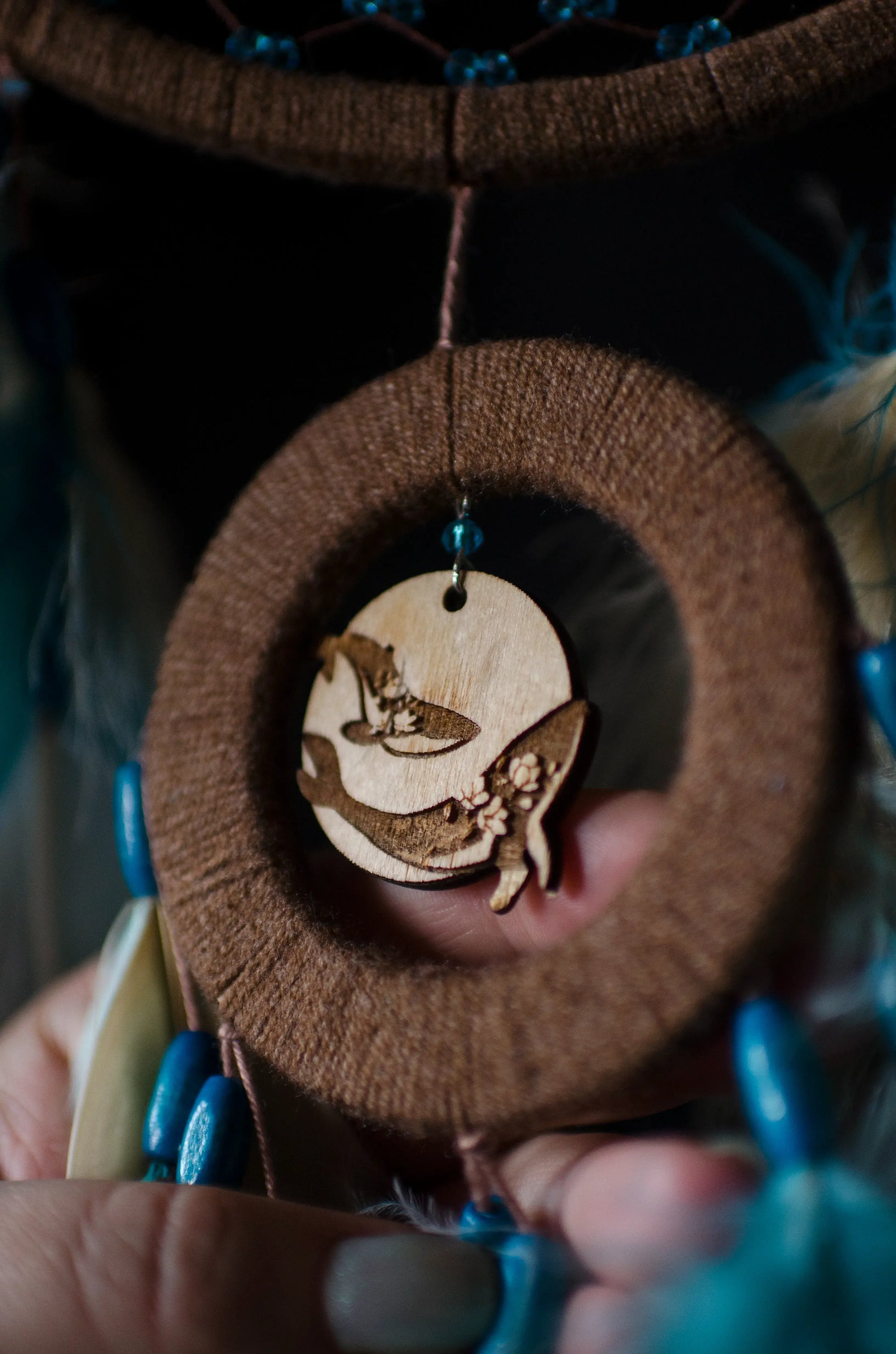 Handcrafted dream catcher with a large brown hoop featuring an intricate web adorned with blue beads. Decorated with natural feathers in shades of blue, brown, and white, it includes a central wooden charm with an engraved design. The dream catcher is set against a backdrop of vintage maps and celestial charts, creating a mystical and adventurous aesthetic. Perfect for adding a bohemian touch to any space, it symbolizes protection and positive energy, making it an ideal gift or home decor piece for dreamers