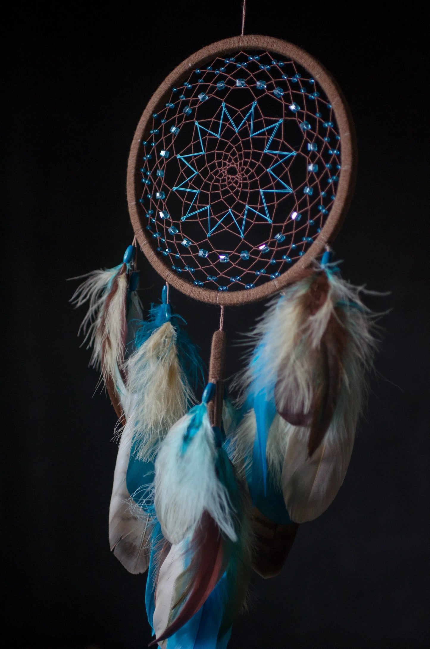 Handcrafted dream catcher with a large brown hoop featuring an intricate web adorned with blue beads. Decorated with natural feathers in shades of blue, brown, and white, it includes a central wooden charm with an engraved design. The dream catcher is set against a backdrop of vintage maps and celestial charts, creating a mystical and adventurous aesthetic. Perfect for adding a bohemian touch to any space, it symbolizes protection and positive energy, making it an ideal gift or home decor piece for dreamers