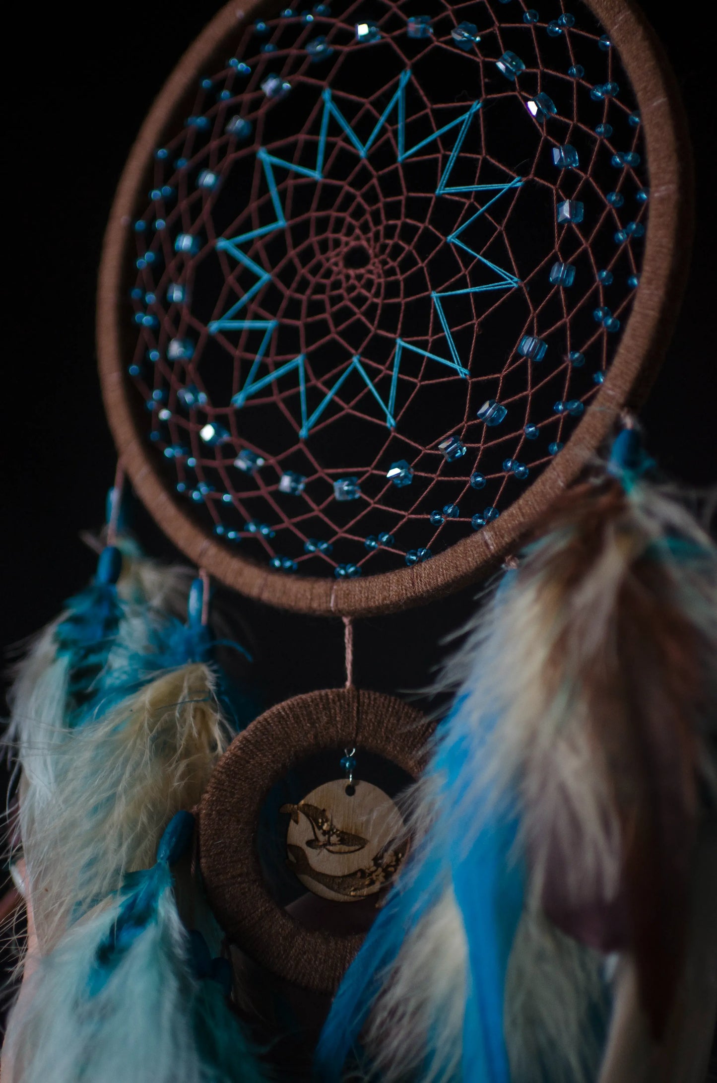 Handcrafted dream catcher with a large brown hoop featuring an intricate web adorned with blue beads. Decorated with natural feathers in shades of blue, brown, and white, it includes a central wooden charm with an engraved design. The dream catcher is set against a backdrop of vintage maps and celestial charts, creating a mystical and adventurous aesthetic. Perfect for adding a bohemian touch to any space, it symbolizes protection and positive energy, making it an ideal gift or home decor piece for dreamers