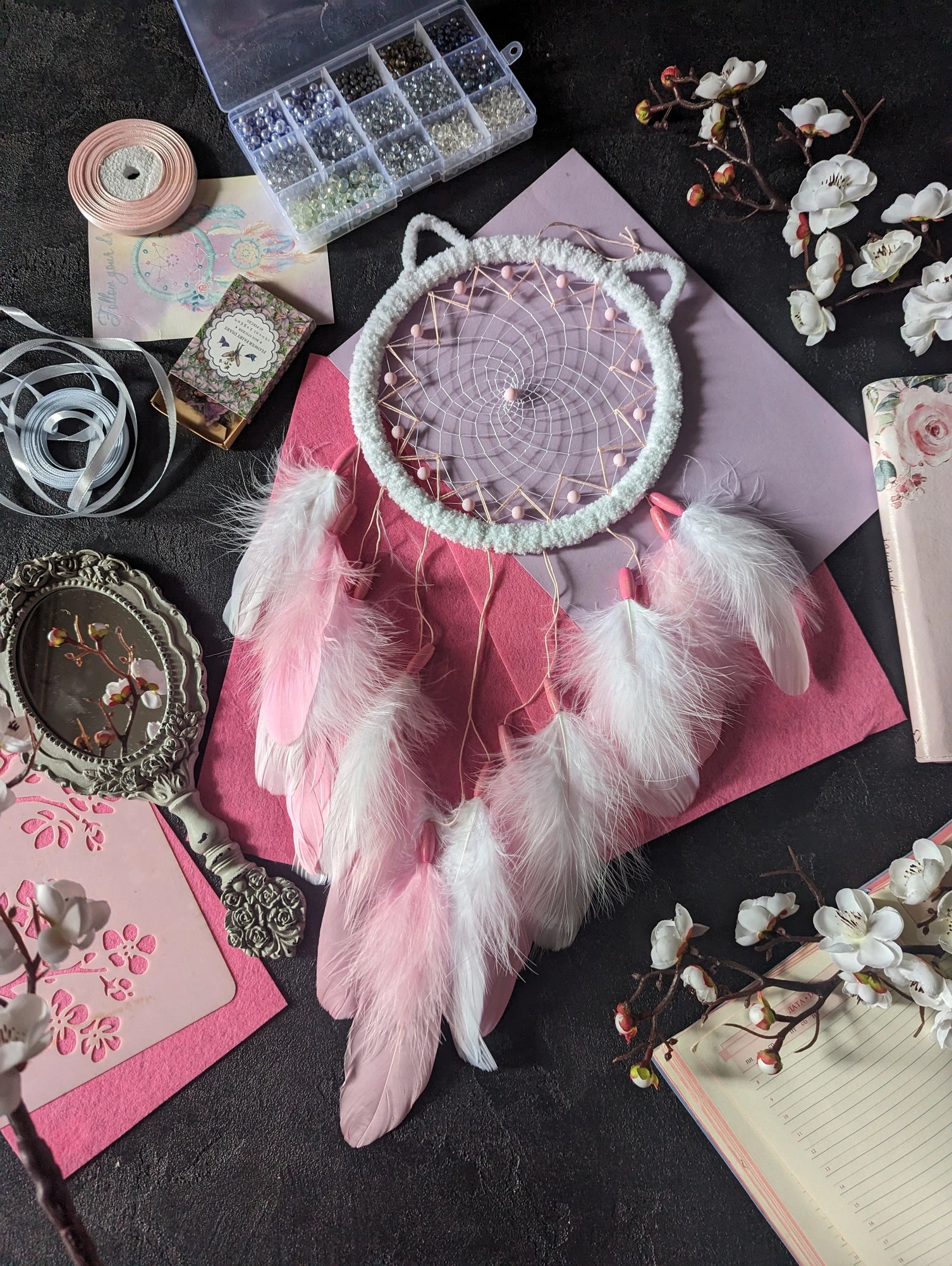 Handcrafted pink and white dream catcher with a cat ear design, featuring a pink beaded web and delicate white and pink feathers. This bohemian decor piece is surrounded by crafting materials and floral elements, making it an ideal gift for a nursery or girl's room. The soft pastel colors and intricate detailing add a touch of whimsy and charm, perfect for hanging in a bedroom or as a unique decorative accent in any space. Enhance your home decor with this adorable, handmade dream catcher.