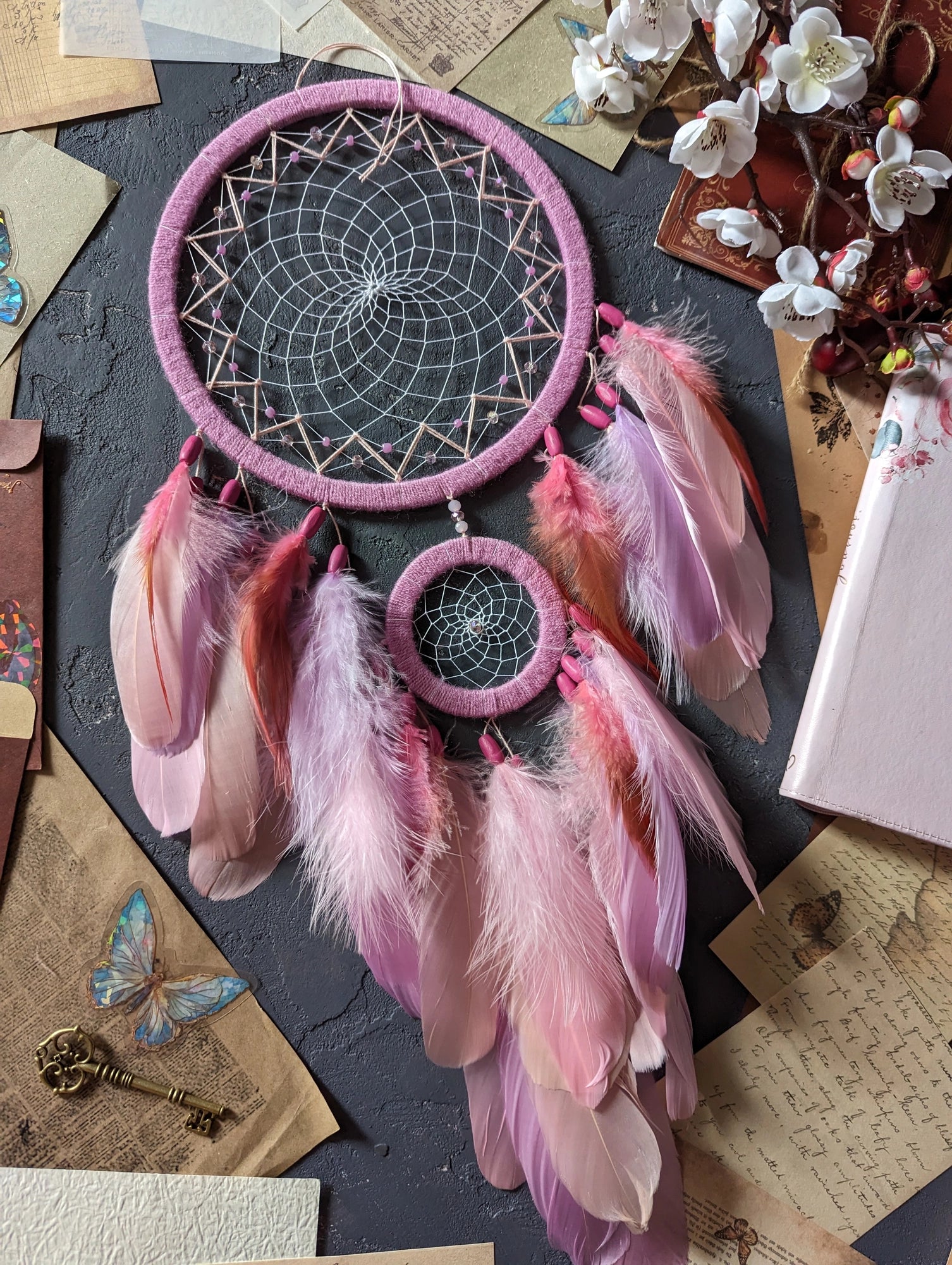 This dream catcher features a large pink hoop with an intricate web adorned with pink beads. Below, it displays an array of soft pink and orange feathers, accented with additional pink beads. The design is complemented by surrounding vintage letters, keys, and delicate cherry blossoms, creating a charming and whimsical aesthetic perfect for any space.