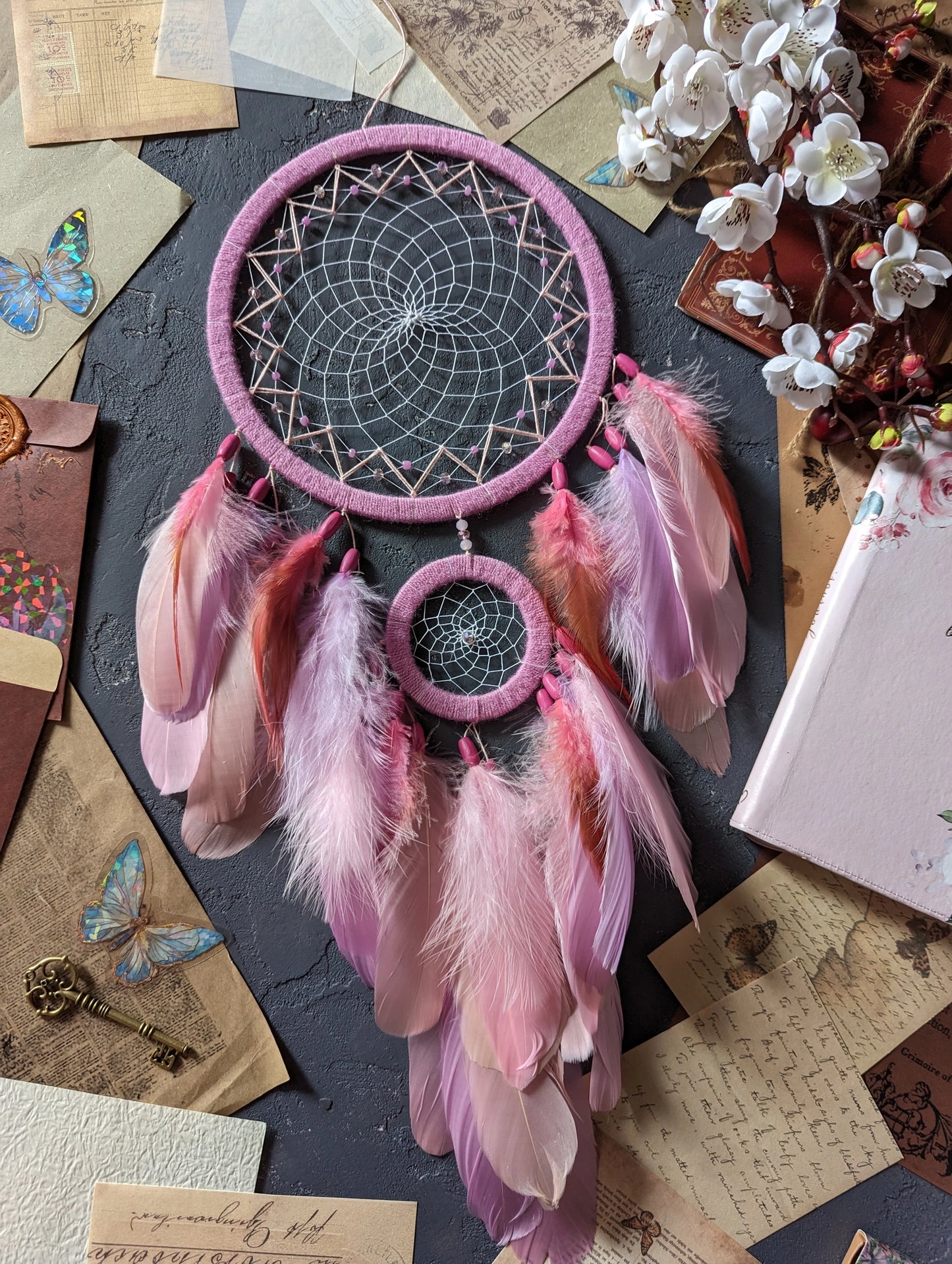 This dream catcher features a large pink hoop with an intricate web adorned with pink beads. Below, it displays an array of soft pink and orange feathers, accented with additional pink beads. The design is complemented by surrounding vintage letters, keys, and delicate cherry blossoms, creating a charming and whimsical aesthetic perfect for any space.