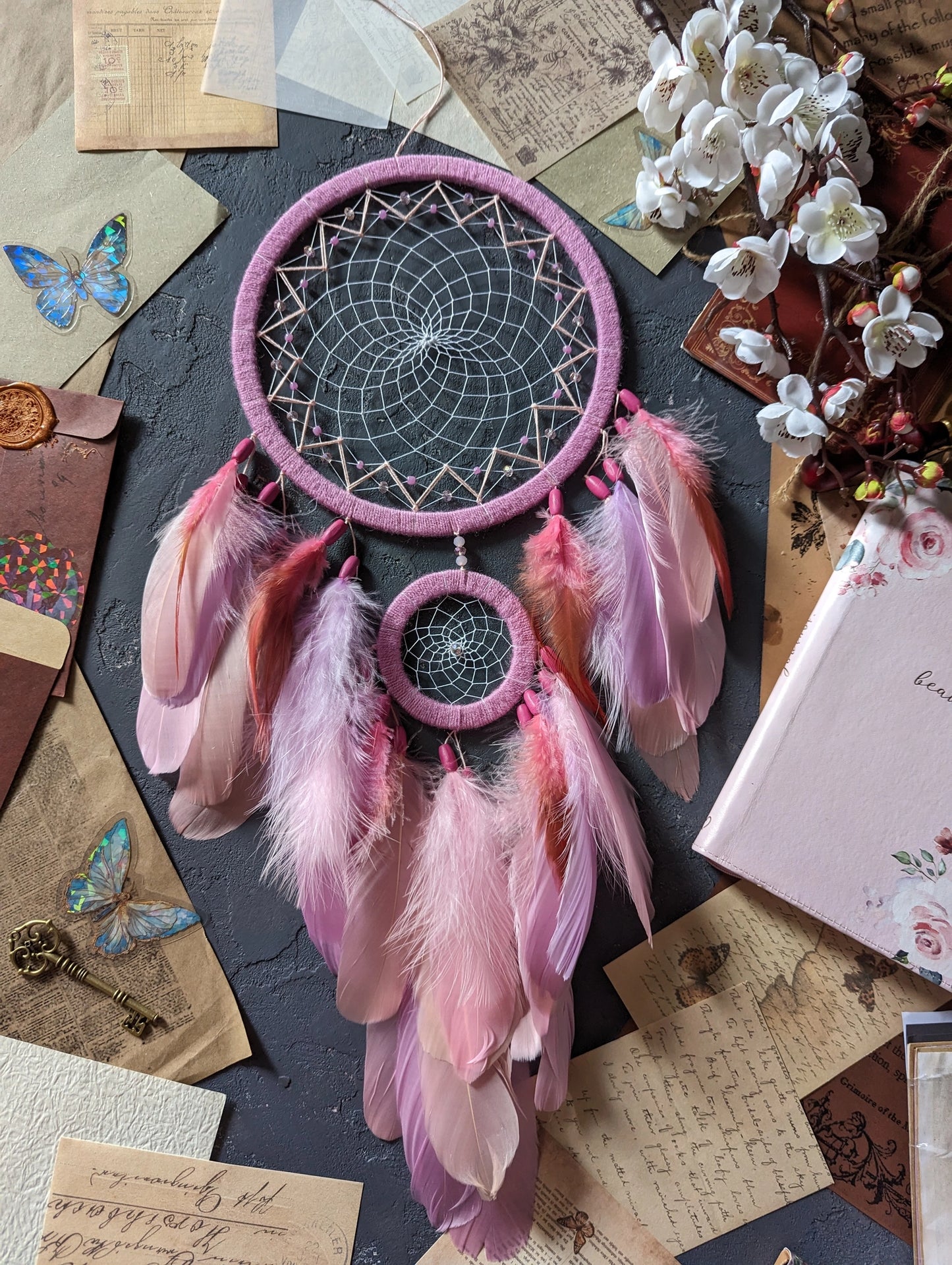 This dream catcher features a large pink hoop with an intricate web adorned with pink beads. Below, it displays an array of soft pink and orange feathers, accented with additional pink beads. The design is complemented by surrounding vintage letters, keys, and delicate cherry blossoms, creating a charming and whimsical aesthetic perfect for any space.