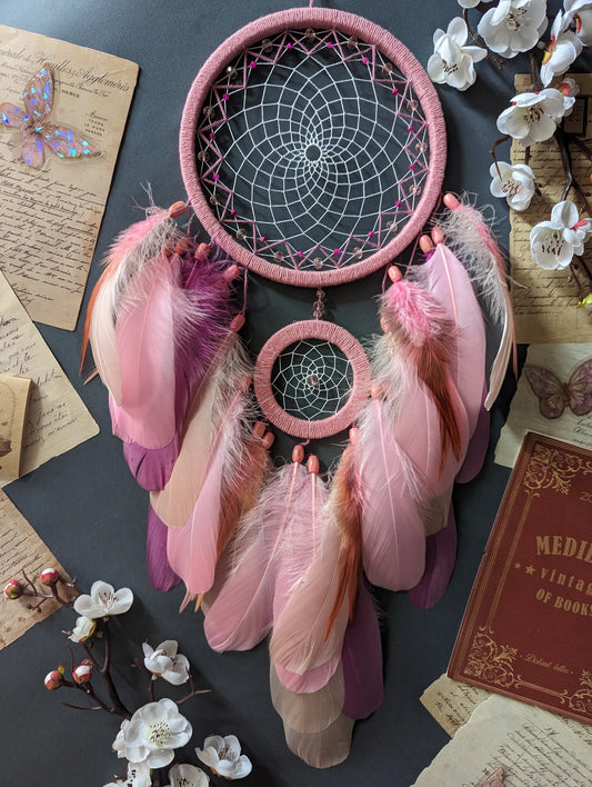 This dream catcher features a large pink hoop with an intricate web adorned with pink beads. Below, it displays an array of soft pink and orange feathers, accented with additional pink beads. The design is complemented by surrounding vintage letters, keys, and delicate cherry blossoms, creating a charming and whimsical aesthetic perfect for any space.