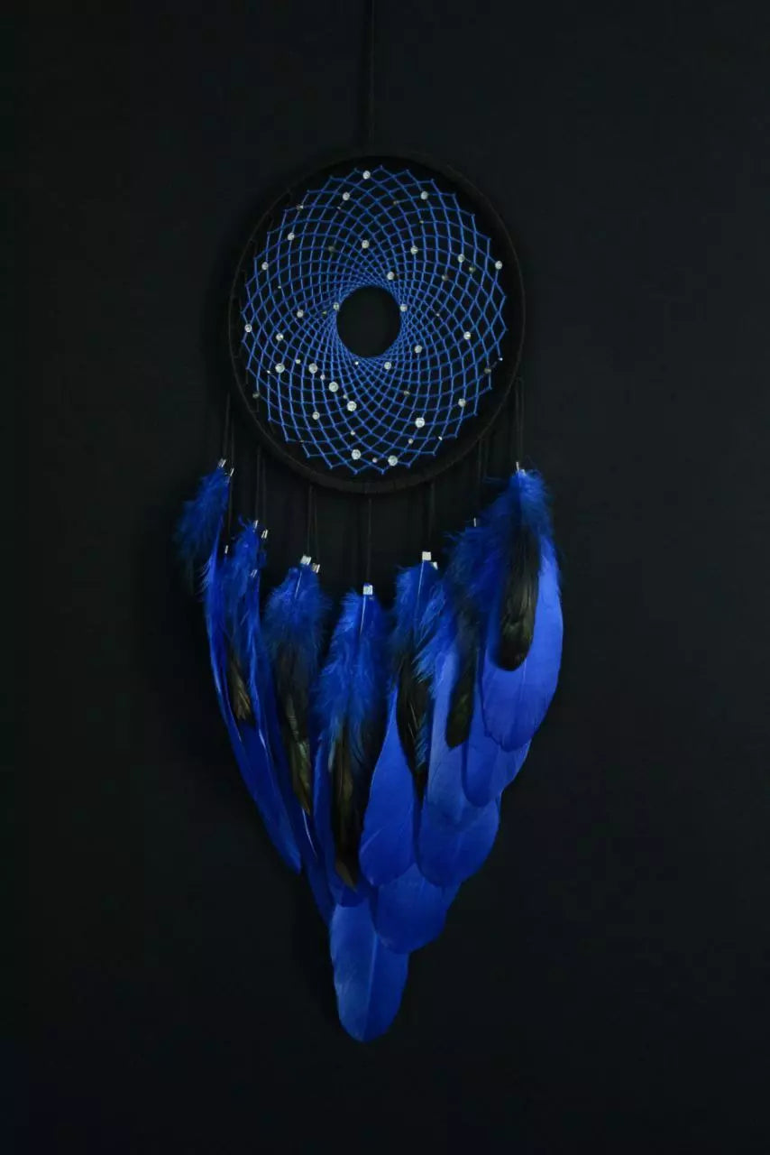 Handmade dreamcatcher with a black hoop, featuring a meticulously woven web adorned with small reflective beads, creating a galaxy-like effect. The dreamcatcher is embellished with vibrant blue and purple feathers, adding a striking contrast and visual appeal. Perfect for adding a touch of mystique and elegance to any space.