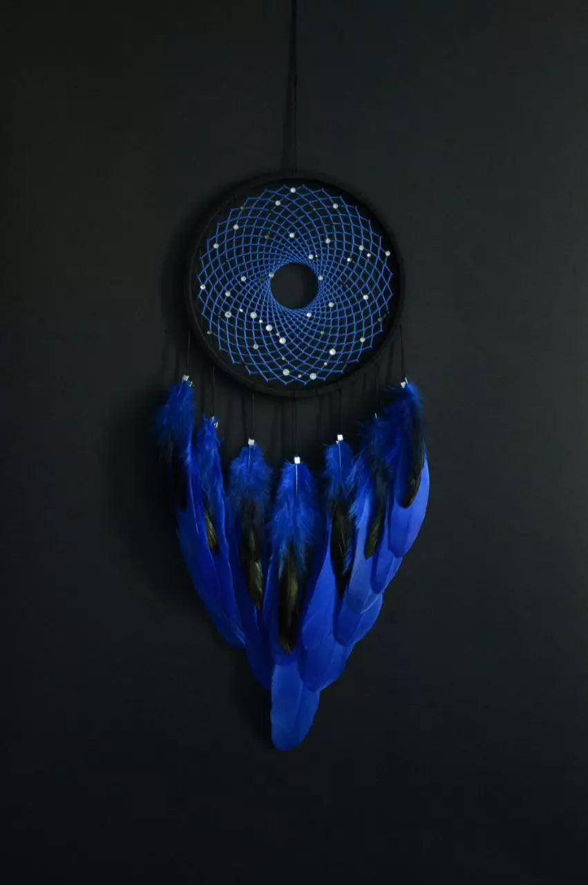 Handmade dreamcatcher with a black hoop, featuring a meticulously woven web adorned with small reflective beads, creating a galaxy-like effect. The dreamcatcher is embellished with vibrant blue and purple feathers, adding a striking contrast and visual appeal. Perfect for adding a touch of mystique and elegance to any space.