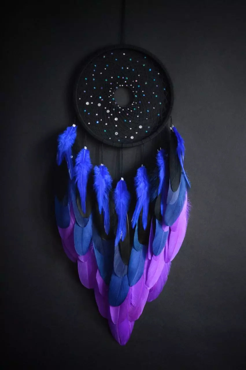 Handmade dreamcatcher with a black hoop, featuring a meticulously woven web adorned with small reflective beads, creating a galaxy-like effect. The dreamcatcher is embellished with vibrant blue and purple feathers, adding a striking contrast and visual appeal. Perfect for adding a touch of mystique and elegance to any space.