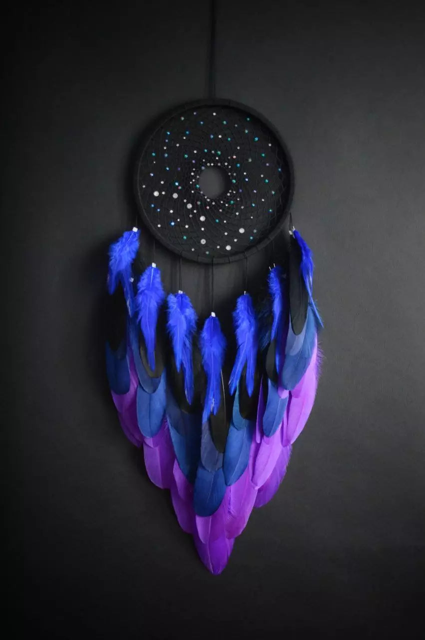 Handmade dreamcatcher with a black hoop, featuring a meticulously woven web adorned with small reflective beads, creating a galaxy-like effect. The dreamcatcher is embellished with vibrant blue and purple feathers, adding a striking contrast and visual appeal. Perfect for adding a touch of mystique and elegance to any space.