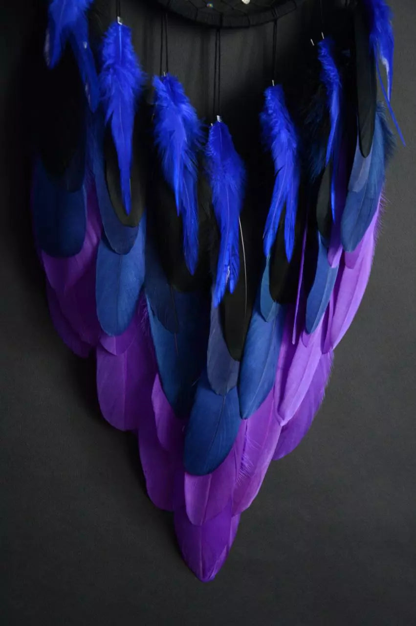 Handmade dreamcatcher with a black hoop, featuring a meticulously woven web adorned with small reflective beads, creating a galaxy-like effect. The dreamcatcher is embellished with vibrant blue and purple feathers, adding a striking contrast and visual appeal. Perfect for adding a touch of mystique and elegance to any space.