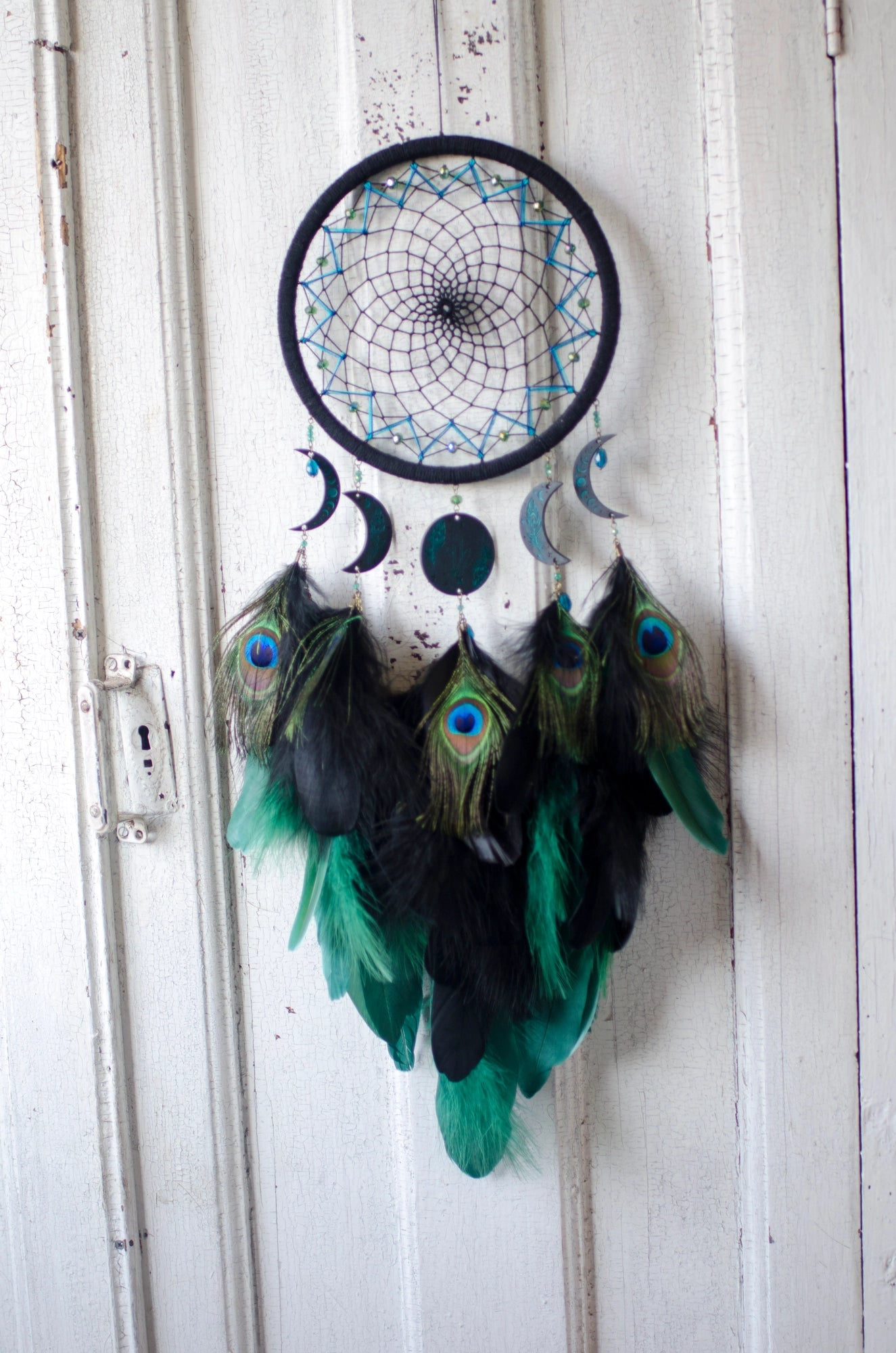 Handcrafted dream catcher featuring an intricate teal web design on a black hoop, adorned with peacock feathers and celestial moon and star charms. This elegant piece is perfect for adding a touch of bohemian flair to any room, symbolizing protection and positive energy.