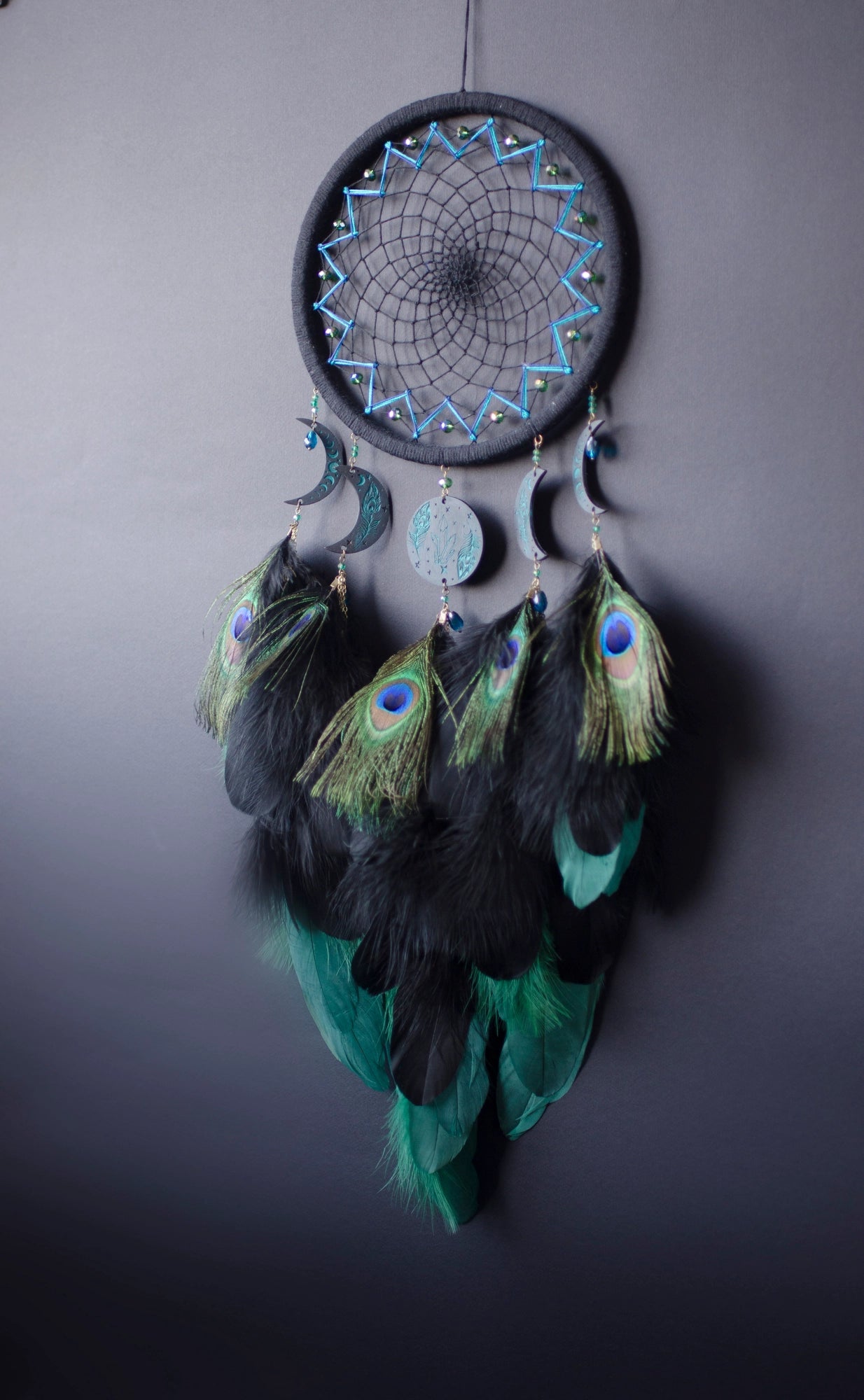 Handcrafted dream catcher featuring an intricate teal web design on a black hoop, adorned with peacock feathers and celestial moon and star charms. This elegant piece is perfect for adding a touch of bohemian flair to any room, symbolizing protection and positive energy.