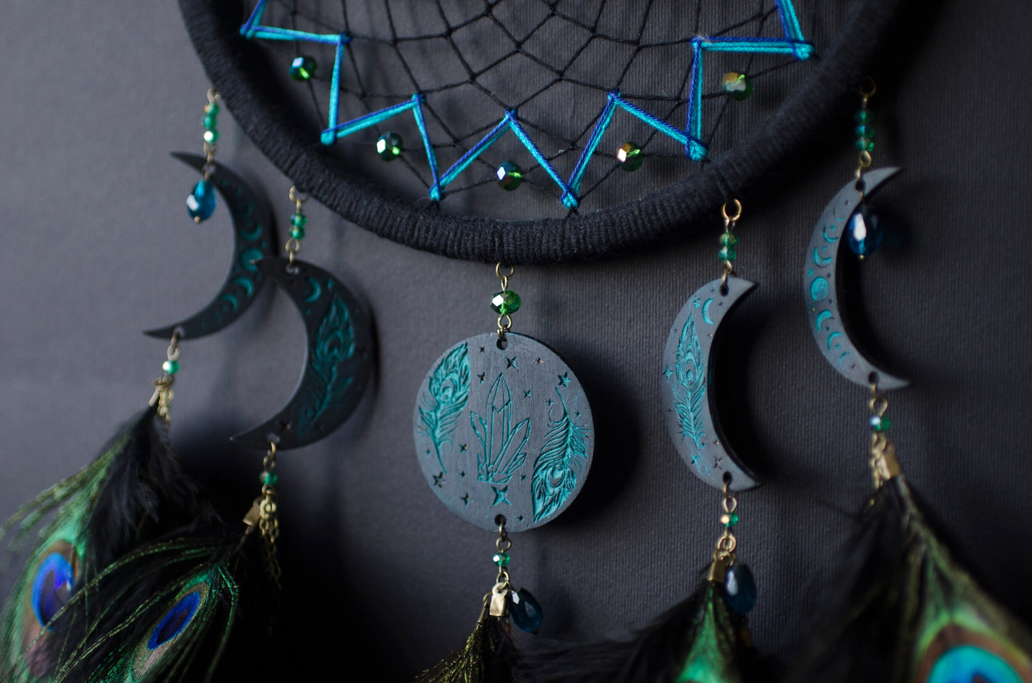 Handcrafted dream catcher featuring an intricate teal web design on a black hoop, adorned with peacock feathers and celestial moon and star charms. This elegant piece is perfect for adding a touch of bohemian flair to any room, symbolizing protection and positive energy.