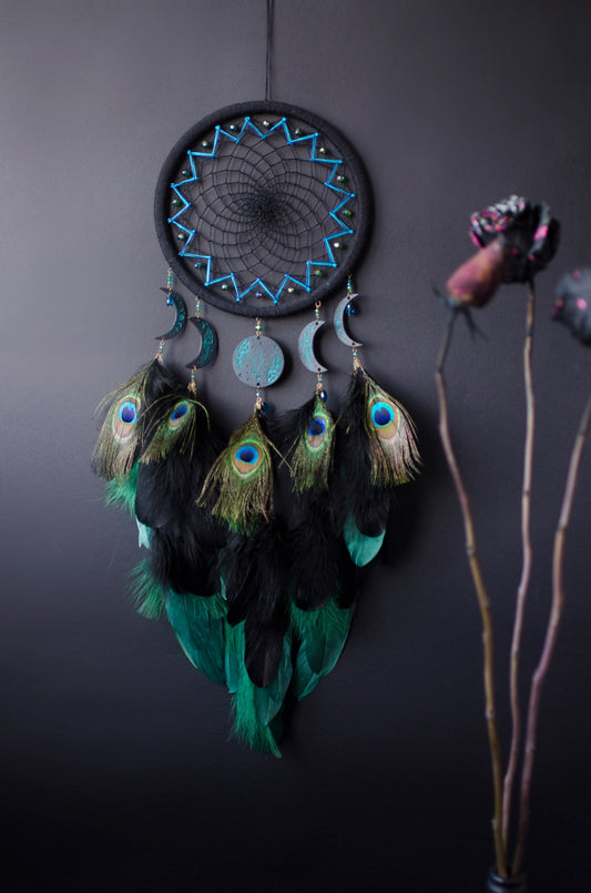 Handcrafted dream catcher featuring an intricate teal web design on a black hoop, adorned with peacock feathers and celestial moon and star charms. This elegant piece is perfect for adding a touch of bohemian flair to any room, symbolizing protection and positive energy.