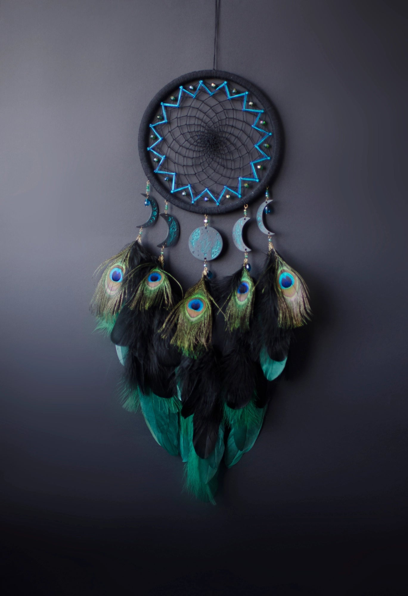 Handcrafted dream catcher featuring an intricate teal web design on a black hoop, adorned with peacock feathers and celestial moon and star charms. This elegant piece is perfect for adding a touch of bohemian flair to any room, symbolizing protection and positive energy.