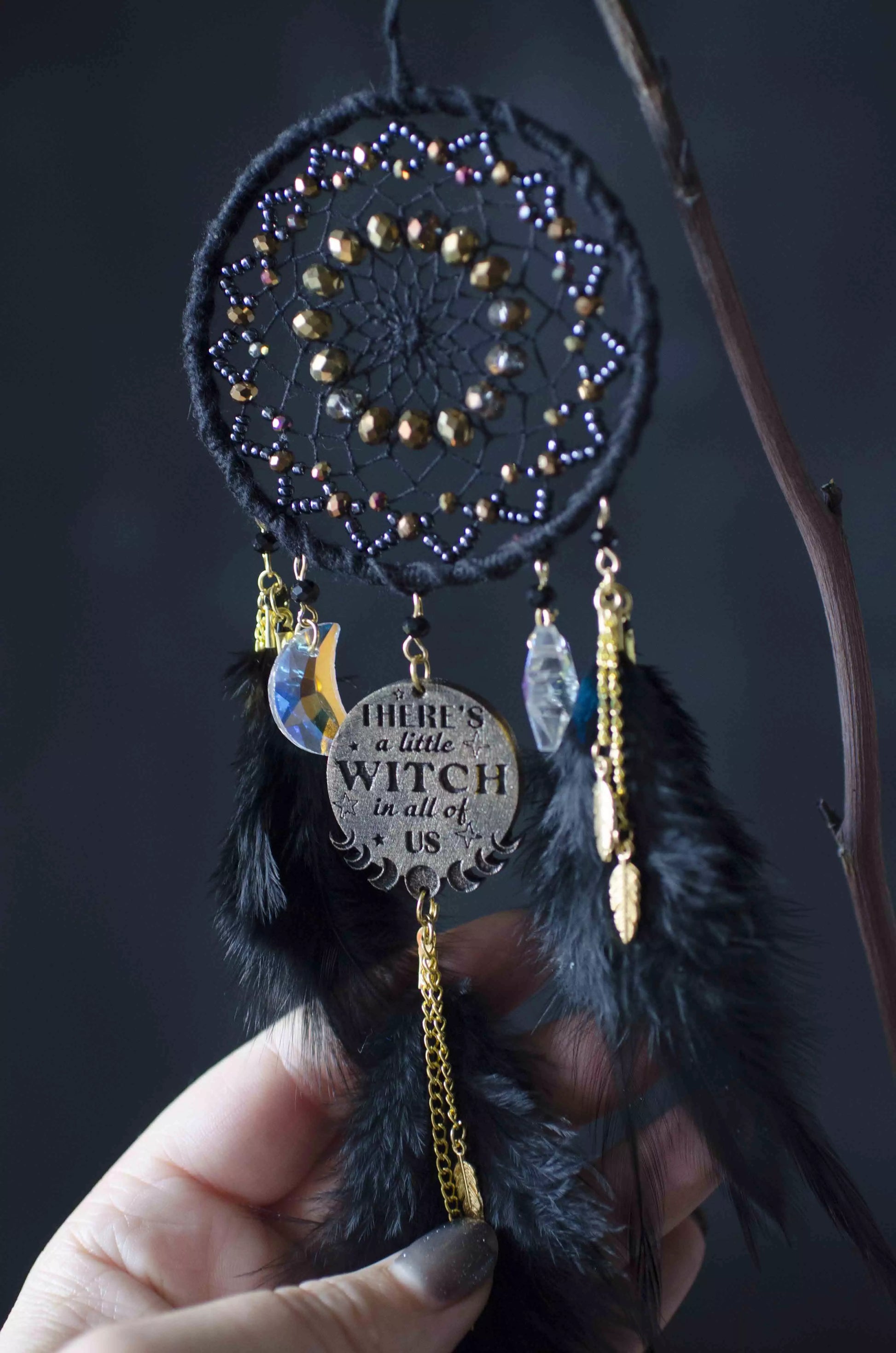 Handcrafted black witch dream catcher featuring a woven black yarn web, golden beads, black feathers, crystal moon and star charms, and a metallic 'There's a little witch in all of us' charm, designed for mystical decor and spiritual protection.