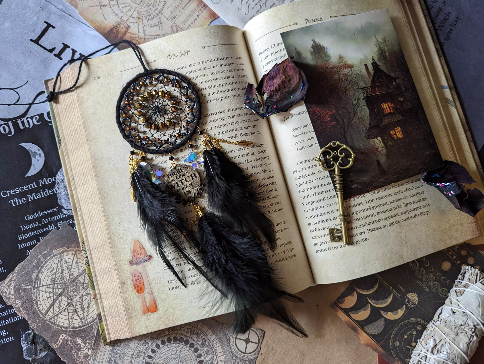 Handcrafted black witch dream catcher featuring a woven black yarn web, golden beads, black feathers, crystal moon and star charms, and a metallic 'There's a little witch in all of us' charm, designed for mystical decor and spiritual protection.