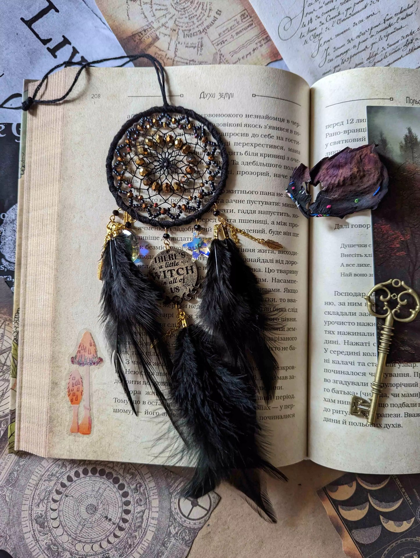 Handcrafted black witch dream catcher featuring a woven black yarn web, golden beads, black feathers, crystal moon and star charms, and a metallic 'There's a little witch in all of us' charm, designed for mystical decor and spiritual protection.