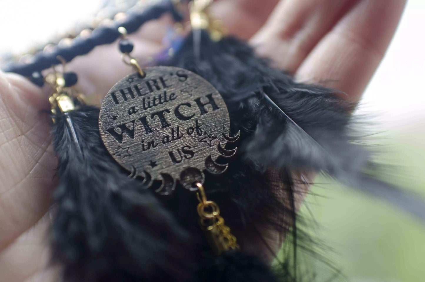 Handcrafted black witch dream catcher featuring a woven black yarn web, golden beads, black feathers, crystal moon and star charms, and a metallic 'There's a little witch in all of us' charm, designed for mystical decor and spiritual protection.