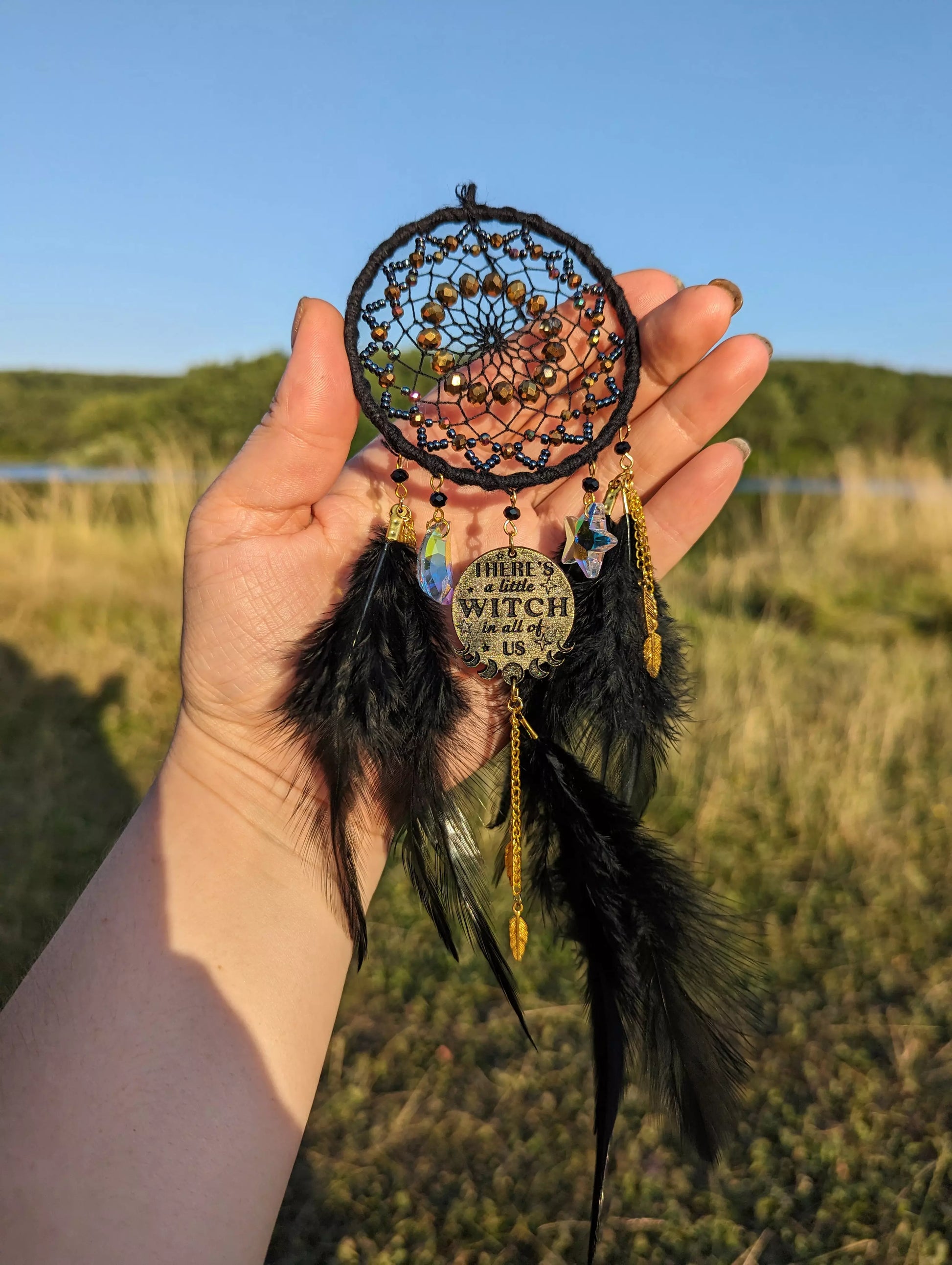 Handcrafted black witch dream catcher featuring a woven black yarn web, golden beads, black feathers, crystal moon and star charms, and a metallic 'There's a little witch in all of us' charm, designed for mystical decor and spiritual protection.