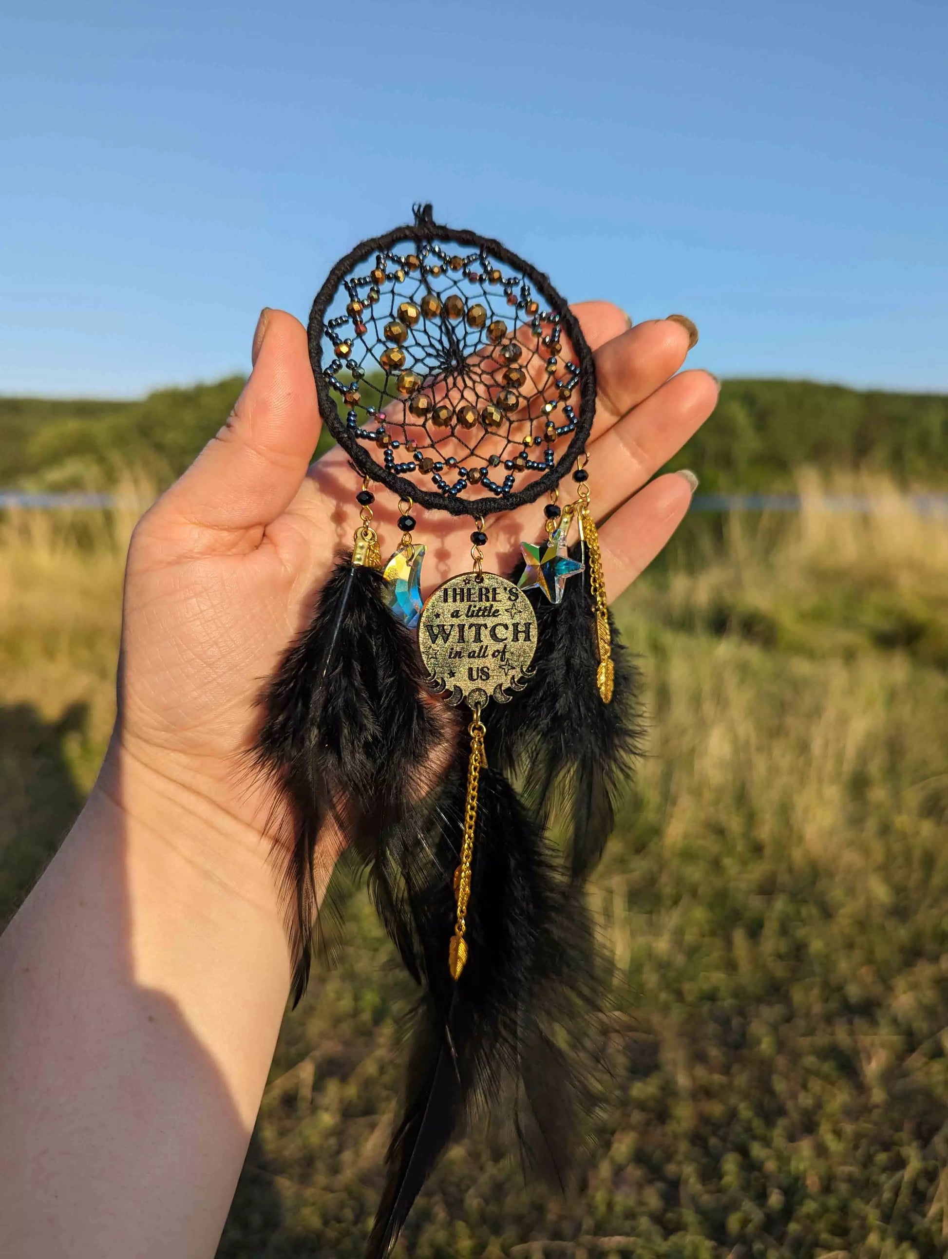 Handcrafted black witch dream catcher featuring a woven black yarn web, golden beads, black feathers, crystal moon and star charms, and a metallic 'There's a little witch in all of us' charm, designed for mystical decor and spiritual protection.