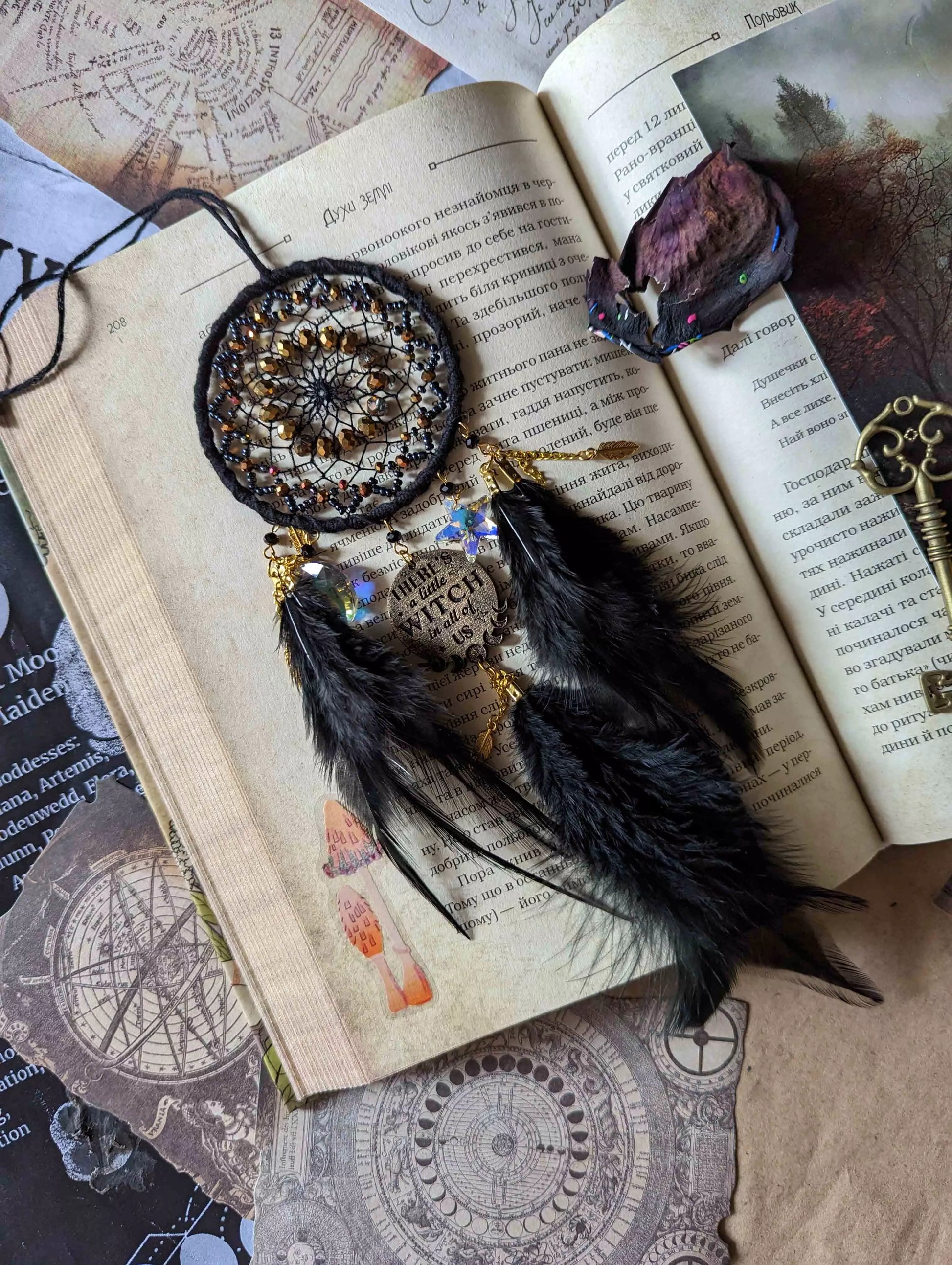 Handcrafted black witch dream catcher featuring a woven black yarn web, golden beads, black feathers, crystal moon and star charms, and a metallic 'There's a little witch in all of us' charm, designed for mystical decor and spiritual protection.