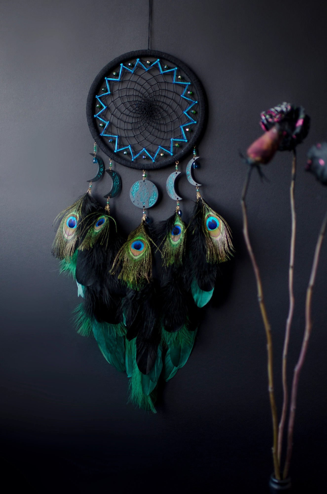 Handcrafted dream catcher featuring an intricate teal web design on a black hoop, adorned with peacock feathers and celestial moon and star charms. This elegant piece is perfect for adding a touch of bohemian flair to any room, symbolizing protection and positive energy.