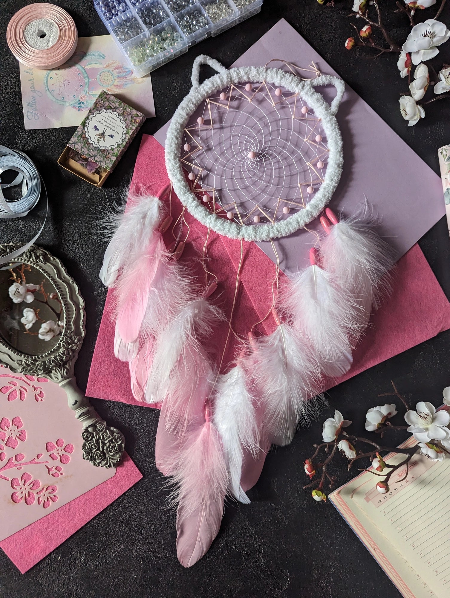 Handcrafted pink and white dream catcher with a cat ear design, featuring a pink beaded web and delicate white and pink feathers. This bohemian decor piece is surrounded by crafting materials and floral elements, making it an ideal gift for a nursery or girl's room. The soft pastel colors and intricate detailing add a touch of whimsy and charm, perfect for hanging in a bedroom or as a unique decorative accent in any space. Enhance your home decor with this adorable, handmade dream catcher.