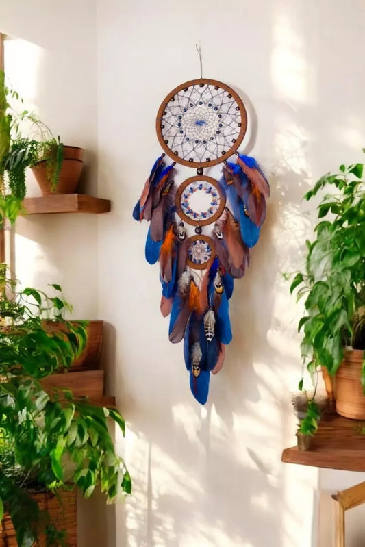 Handcrafted triple hoop dreamcatcher with brown wooden rings, blue and brown feathers, and beaded web, hanging on a wall surrounded by indoor plants in natural sunlight.