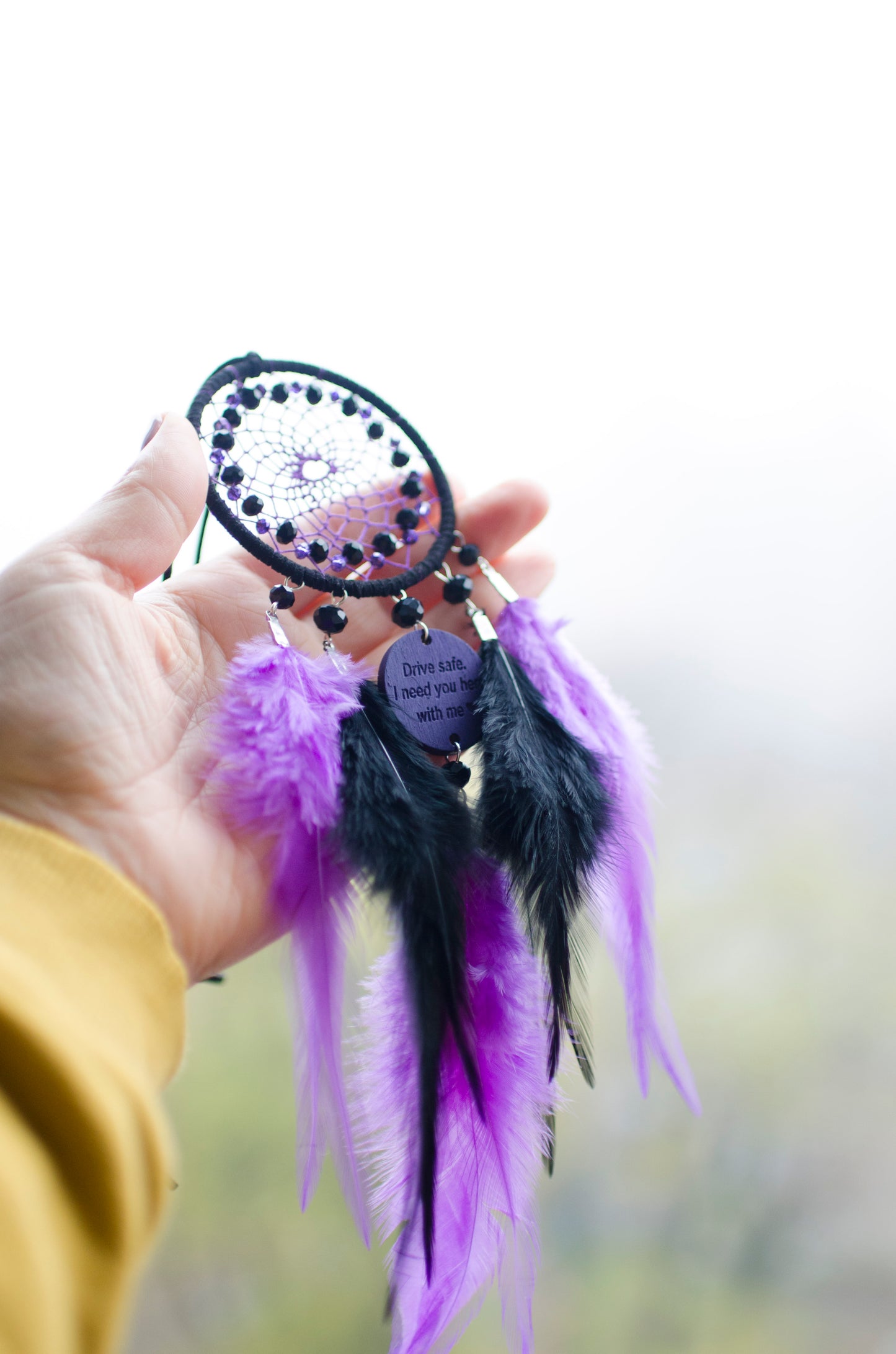 Handcrafted purple and black dream catcher with beaded web design, adorned with vibrant feathers. The centerpiece includes a charm with an engraved message, making it a thoughtful gift or car accessory.