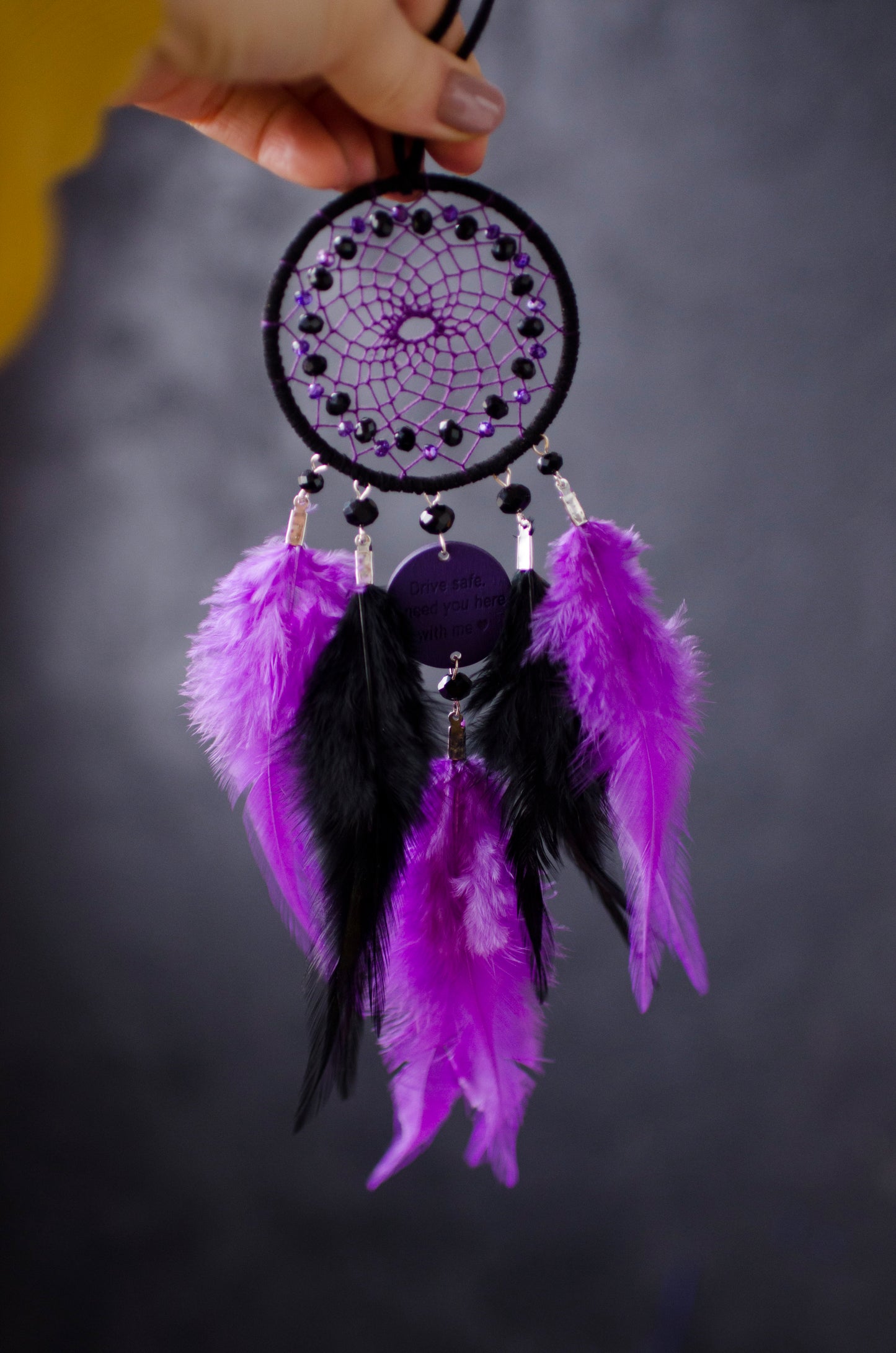 Handcrafted purple and black dream catcher with beaded web design, adorned with vibrant feathers. The centerpiece includes a charm with an engraved message, making it a thoughtful gift or car accessory.