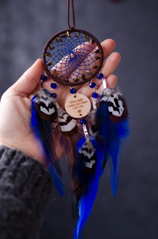 Handcrafted dream catcher featuring a brown hoop with an intricate blue web design. Adorned with tiger's eye beads, vibrant blue crystals, and natural brown and blue feathers. The central wooden charm is engraved with the message, "Drive safe, I need you here with me," making it a perfect talisman for car rearview mirrors. This bohemian-style dream catcher brings positive energy and protection, ideal as a meaningful gift for loved ones or a charming decor piece for cars, homes, or offices.