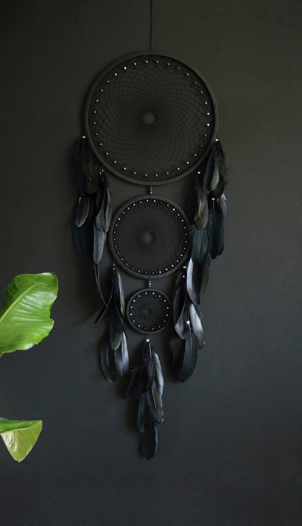 Handcrafted dream catcher featuring a striking black design, with three concentric hoops of varying sizes adorned with intricate webbing. Each hoop is decorated with black beads and complemented by an array of natural black feathers. This elegant piece measures a total length that ensures it becomes a captivating focal point in any room. Perfect for adding a touch of sophistication and positive energy to your home decor. Ideal for those who appreciate unique, artisan-made creations.