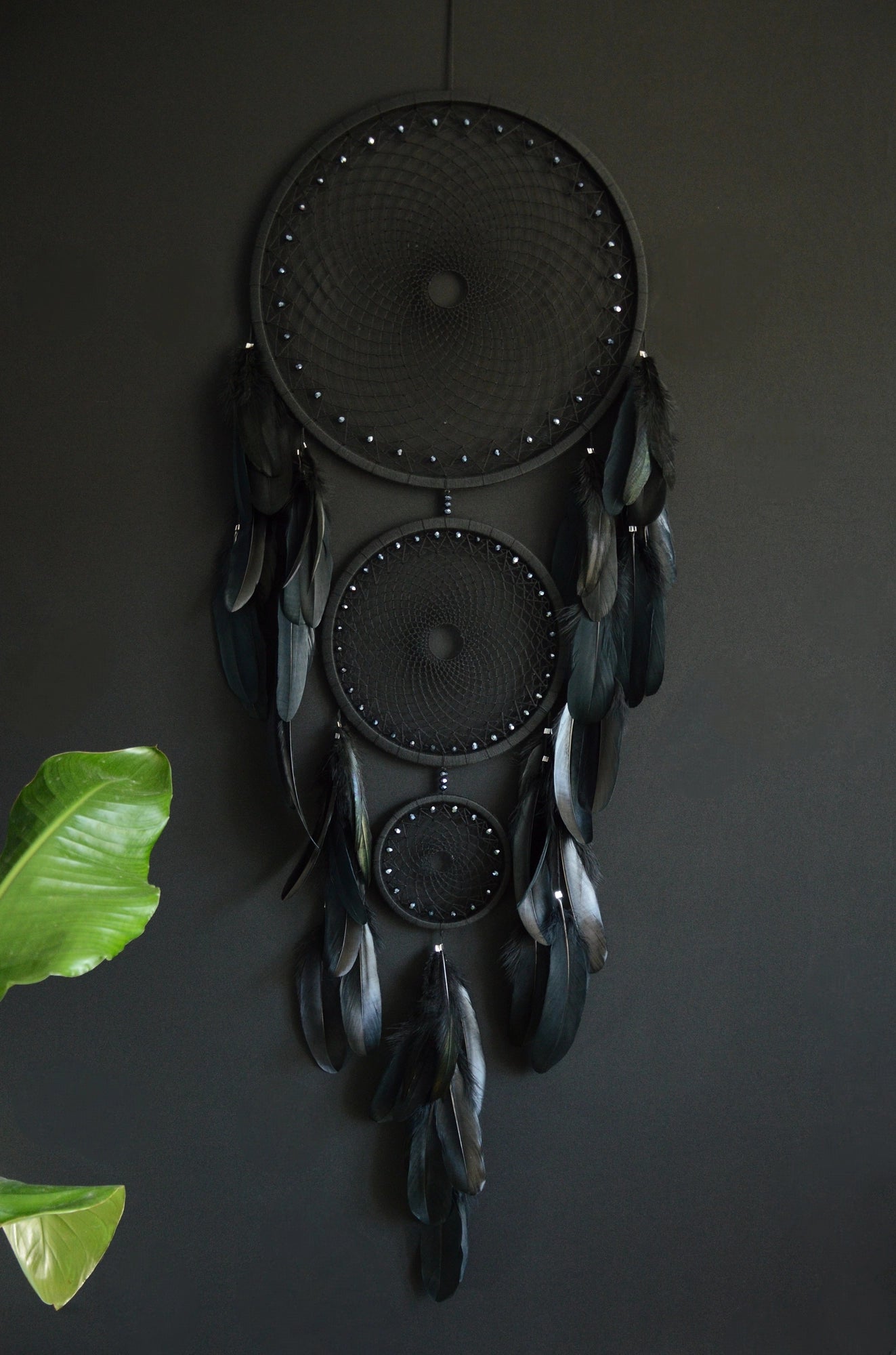 Handcrafted dream catcher featuring a striking black design, with three concentric hoops of varying sizes adorned with intricate webbing. Each hoop is decorated with black beads and complemented by an array of natural black feathers. This elegant piece measures a total length that ensures it becomes a captivating focal point in any room. Perfect for adding a touch of sophistication and positive energy to your home decor. Ideal for those who appreciate unique, artisan-made creations.