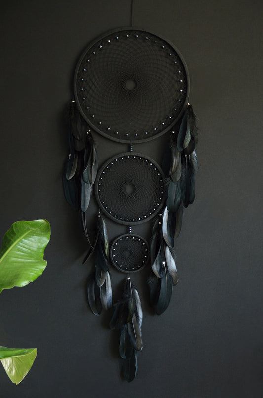 Handcrafted dream catcher featuring a striking black design, with three concentric hoops of varying sizes adorned with intricate webbing. Each hoop is decorated with black beads and complemented by an array of natural black feathers. This elegant piece measures a total length that ensures it becomes a captivating focal point in any room. Perfect for adding a touch of sophistication and positive energy to your home decor. Ideal for those who appreciate unique, artisan-made creations.