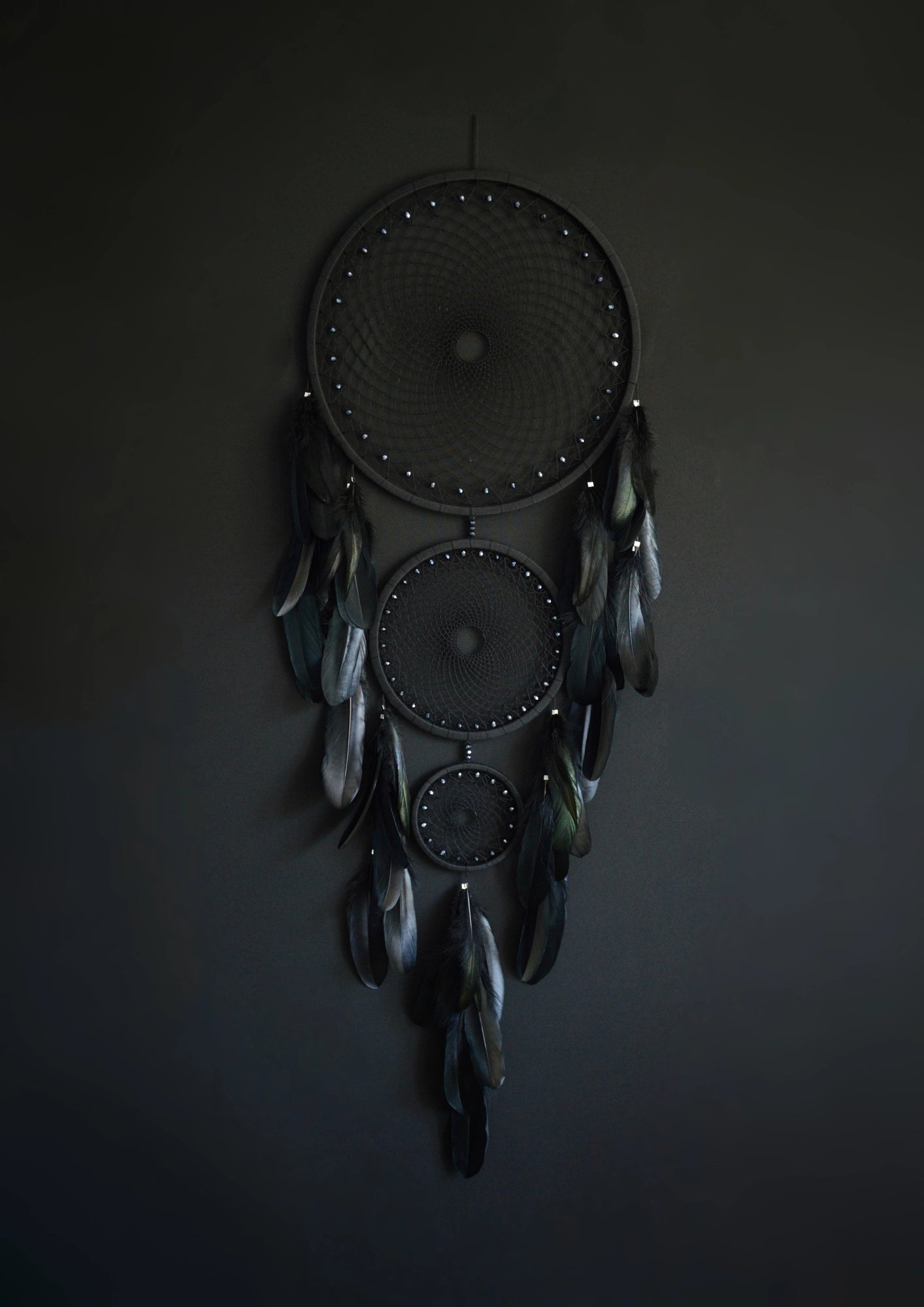 Handcrafted dream catcher featuring a striking black design, with three concentric hoops of varying sizes adorned with intricate webbing. Each hoop is decorated with black beads and complemented by an array of natural black feathers. This elegant piece measures a total length that ensures it becomes a captivating focal point in any room. Perfect for adding a touch of sophistication and positive energy to your home decor. Ideal for those who appreciate unique, artisan-made creations.