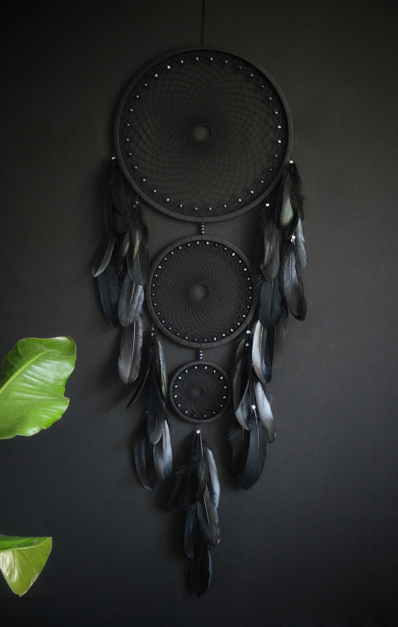 Handcrafted dream catcher featuring a striking black design, with three concentric hoops of varying sizes adorned with intricate webbing. Each hoop is decorated with black beads and complemented by an array of natural black feathers. This elegant piece measures a total length that ensures it becomes a captivating focal point in any room. Perfect for adding a touch of sophistication and positive energy to your home decor. Ideal for those who appreciate unique, artisan-made creations.