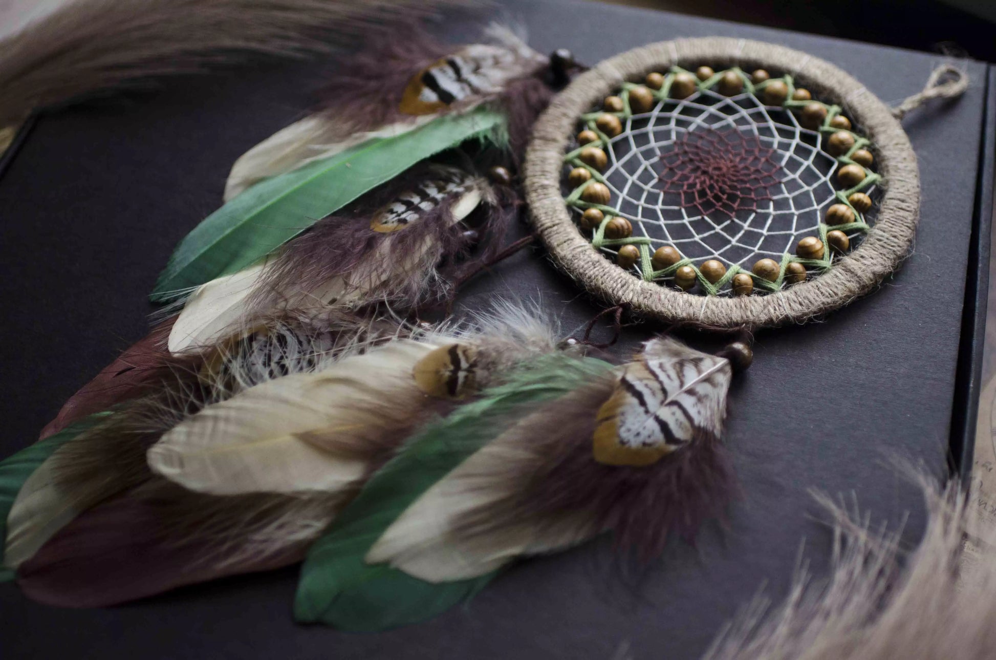 Handmade dream catcher with a 6-inch hoop wrapped in natural jute twine, featuring an intricate web design with wooden beads. Adorned with an array of feathers in green, cream, and brown tones, it serves as a beautiful boho wall decor piece. Perfect for adding a touch of rustic charm to any room.