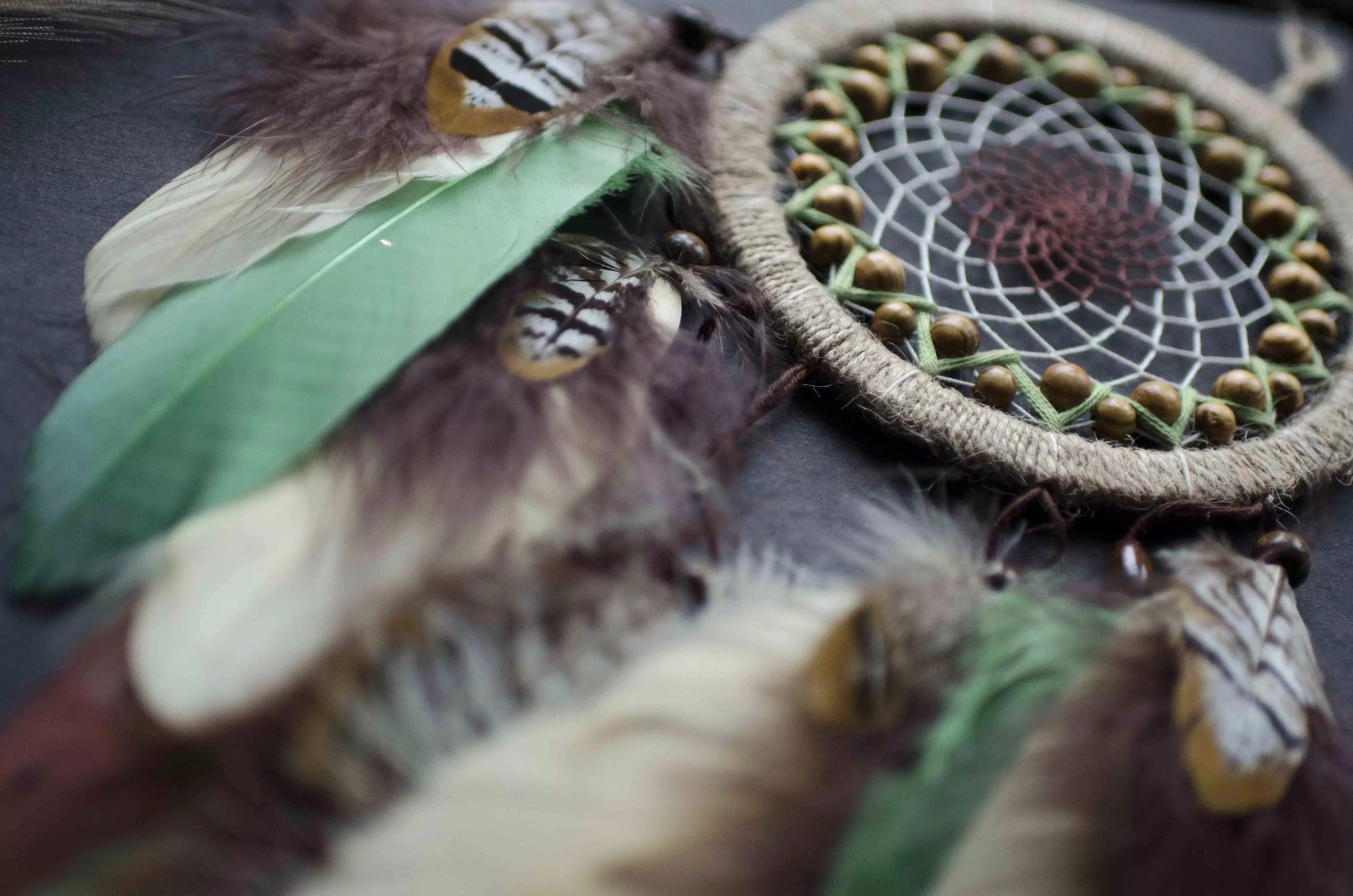 Handmade dream catcher with a 6-inch hoop wrapped in natural jute twine, featuring an intricate web design with wooden beads. Adorned with an array of feathers in green, cream, and brown tones, it serves as a beautiful boho wall decor piece. Perfect for adding a touch of rustic charm to any room.